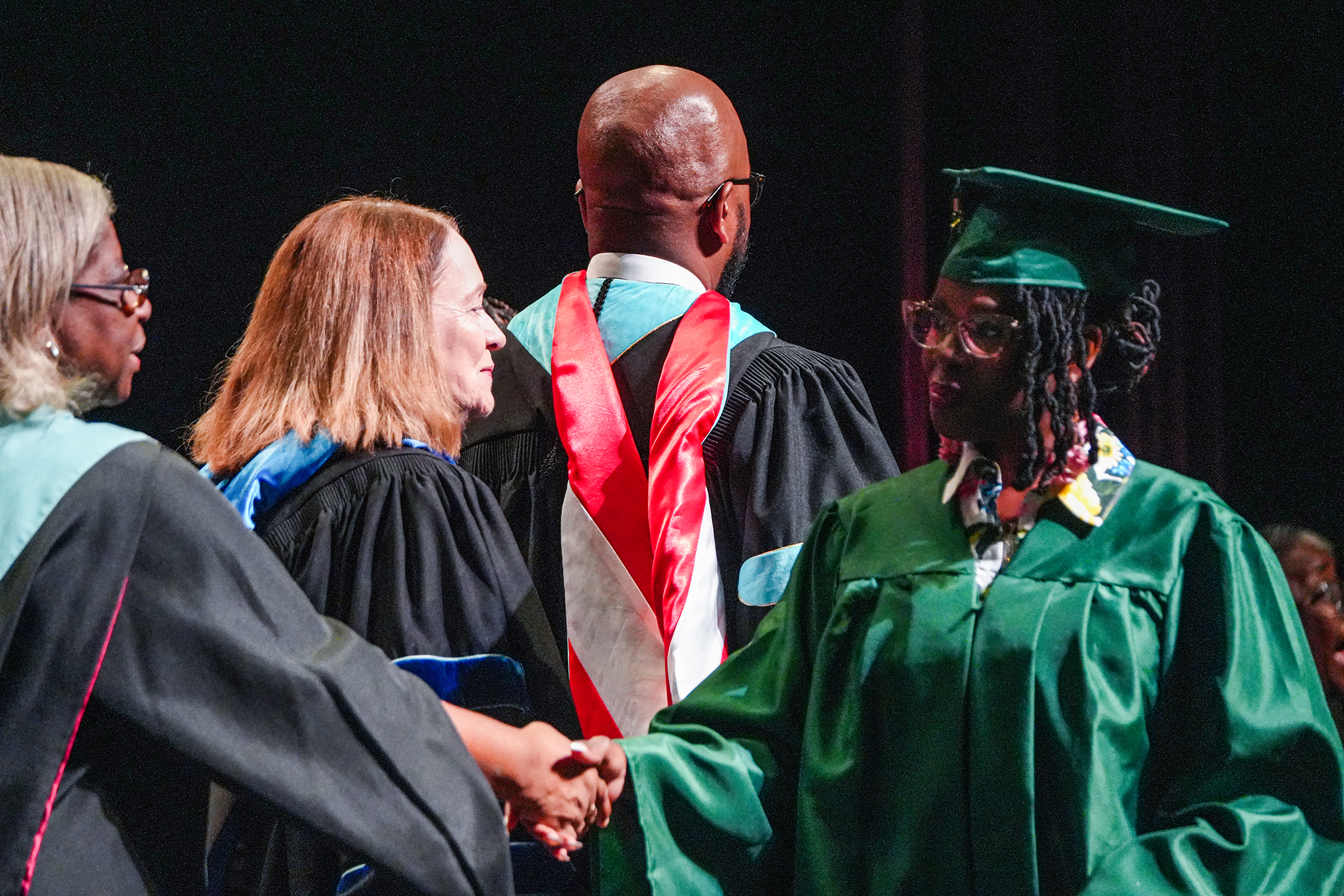 A person in a green graduation gown shakes hands with two others in black gowns. Behind them, a man in a red and white academic gown looks on.
