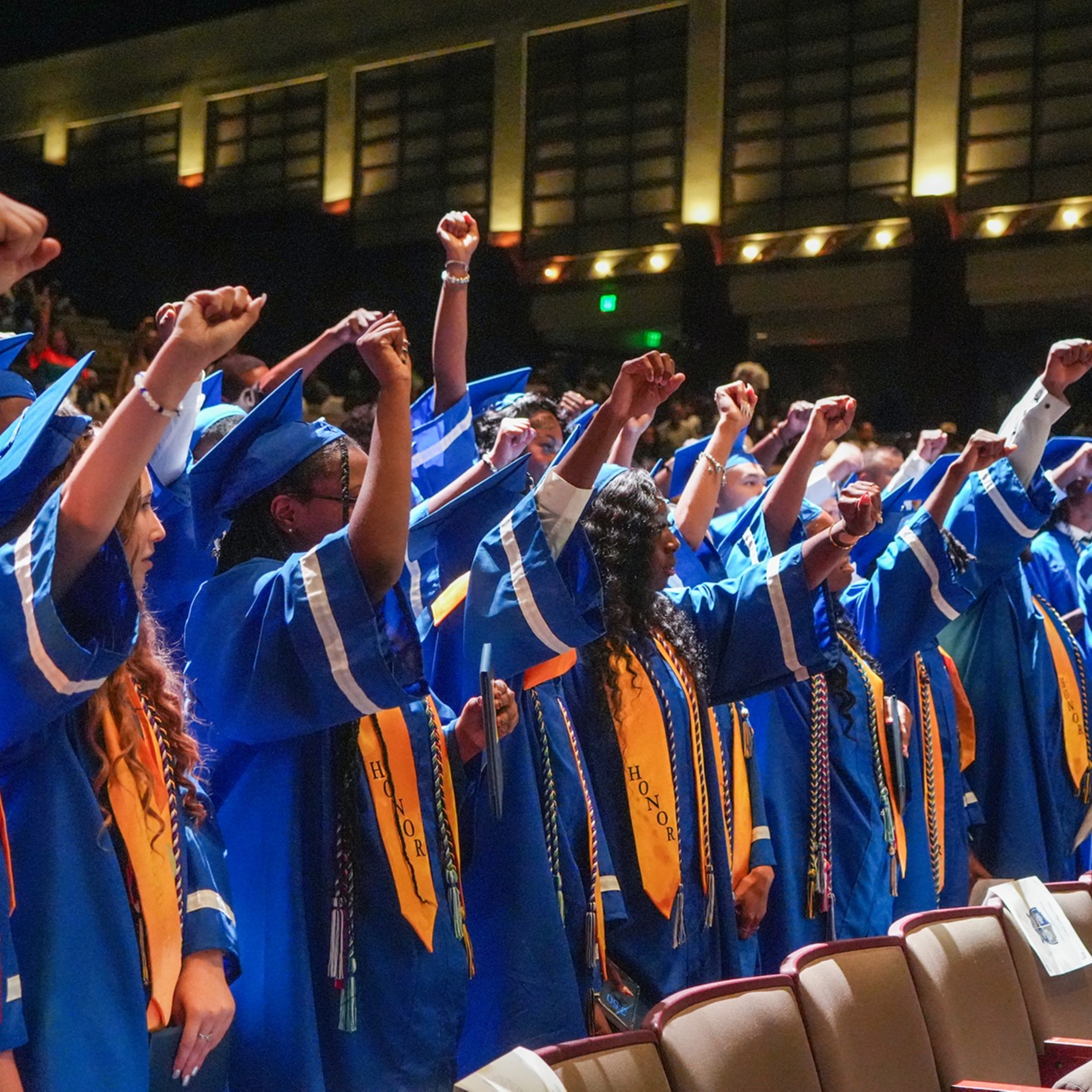 Graduates in blue gowns and caps stand in rows, raising their fists in a unified gesture. The background features a theater with empty seats.