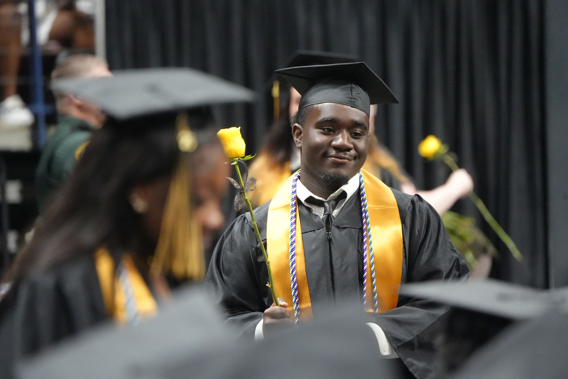 A man in a cap and gown holds a yellow flower at a graduation ceremony with others in caps and gowns.