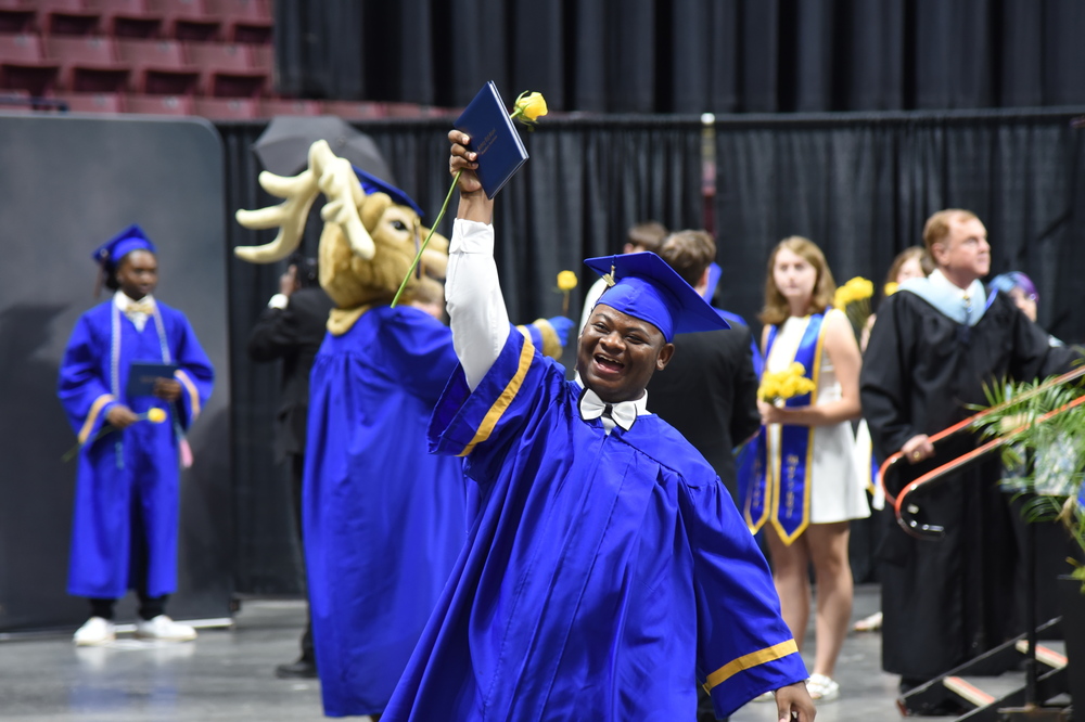A person in a blue cap and gown celebrates, holding a book above their head, with others in similar attire behind them.
