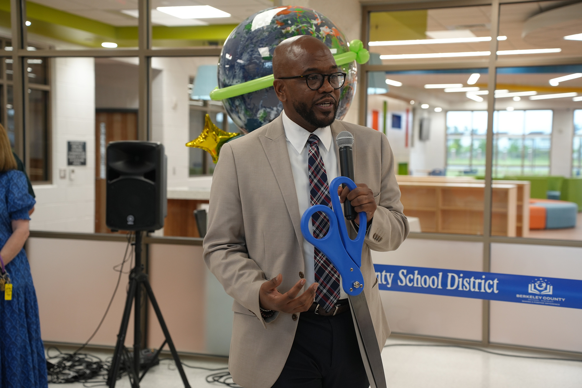 A man in a tan suit and tie holds large blue scissors and a microphone.