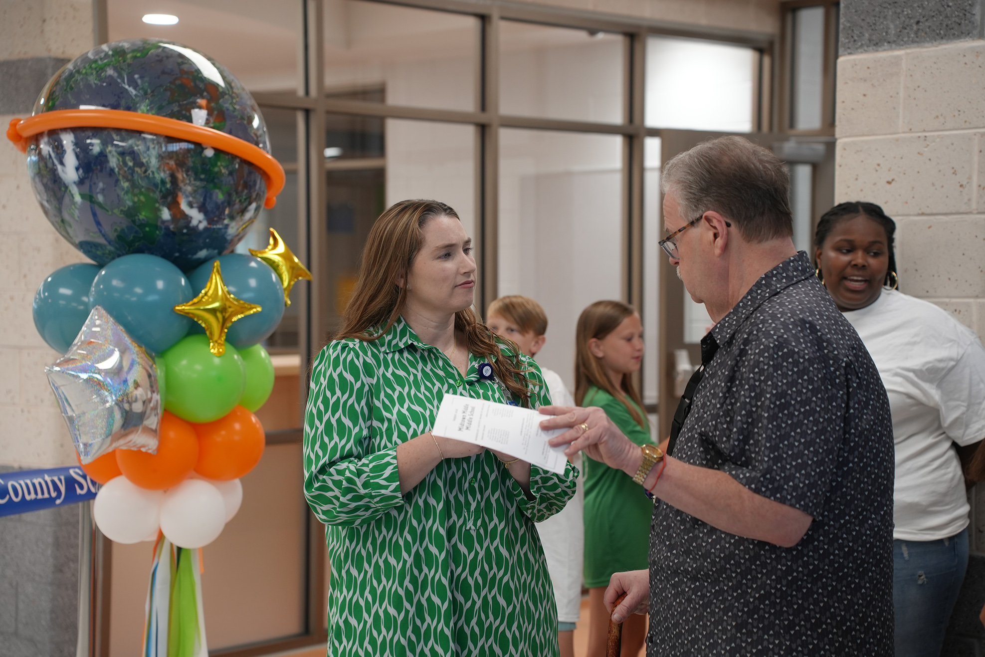 Woman in green dress hands paper to man in gray shirt, balloons in background.
