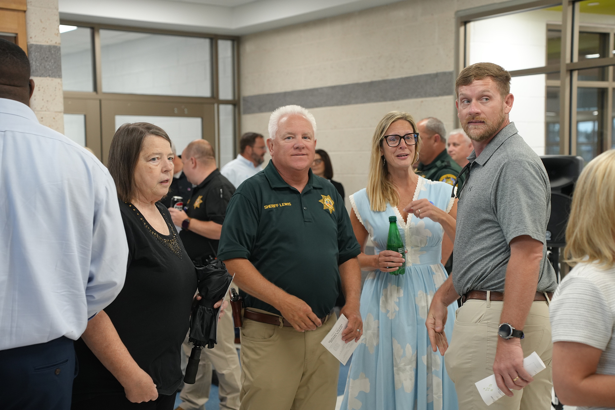Several people stand in a room, including a man in a green uniform, a woman in a white shirt, and others.