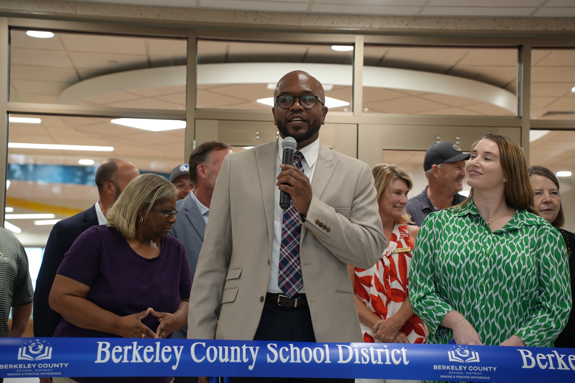 A person holding a microphone speaks at an event with a blue ribbon. Behind them are adults standing, some wearing hats and glasses.
