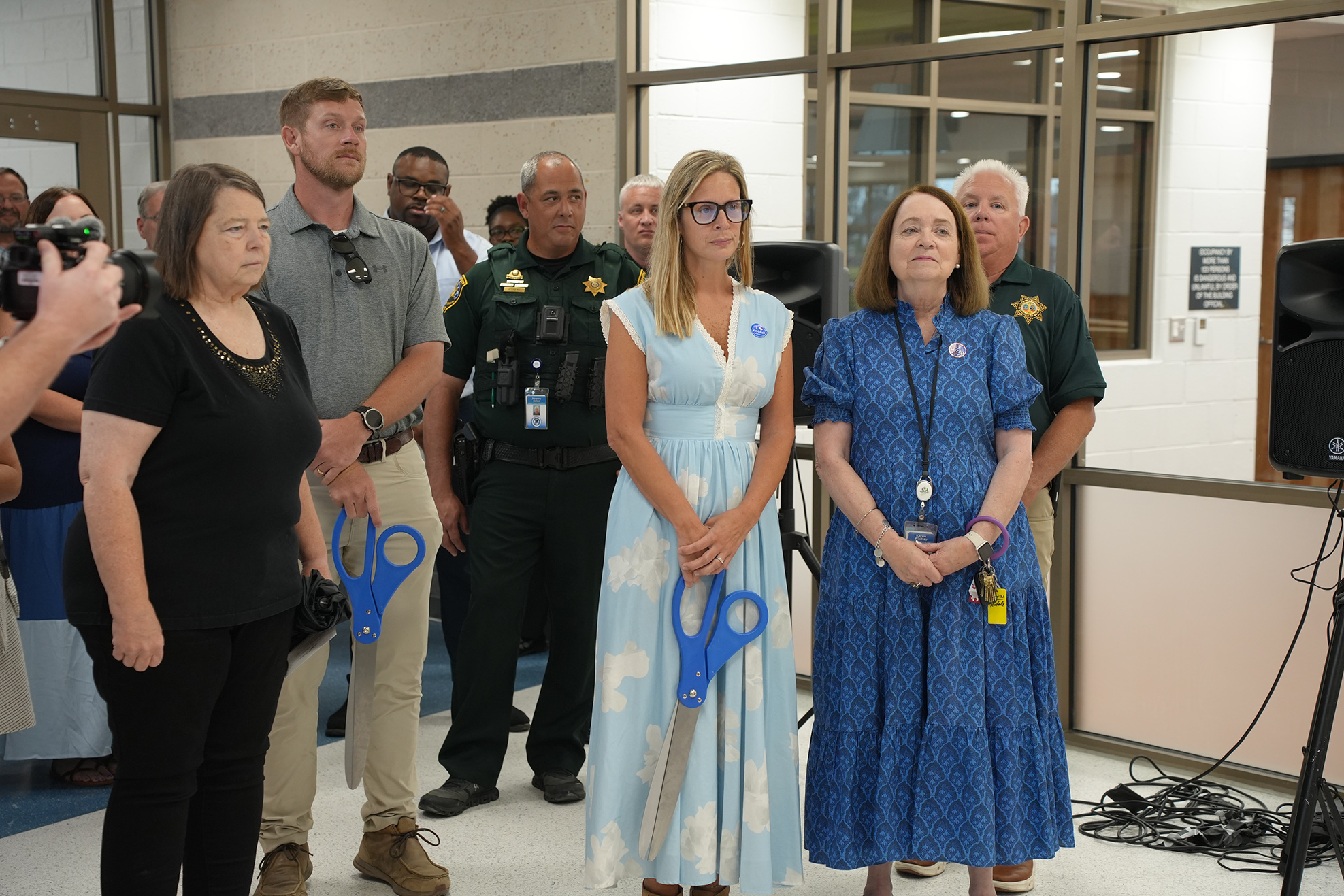 A group of people stand in a room. Three women hold scissors in front of them. Two men in uniforms stand behind them.