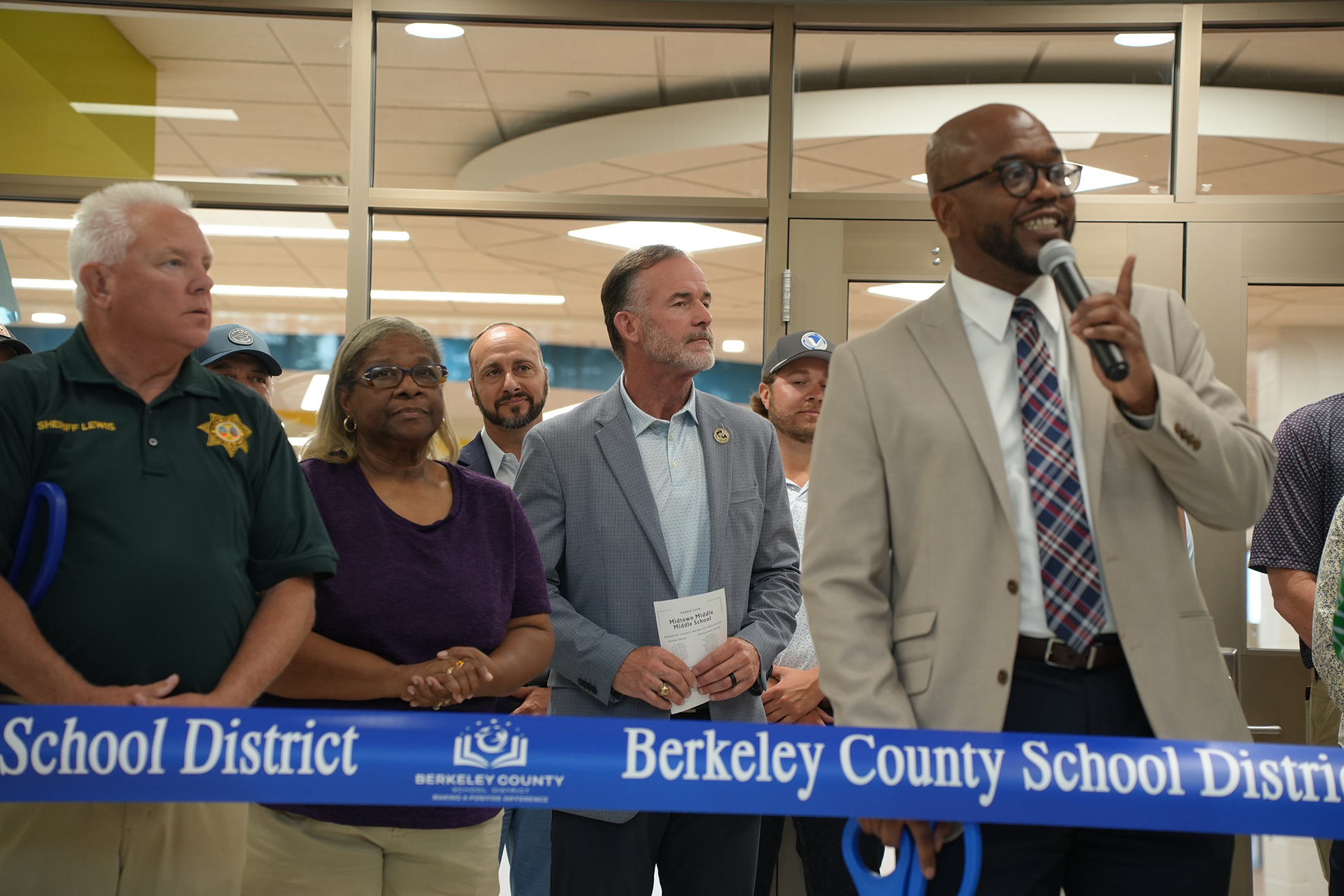 Group of people stand behind a blue ribbon. A man holds a microphone and appears to speak.