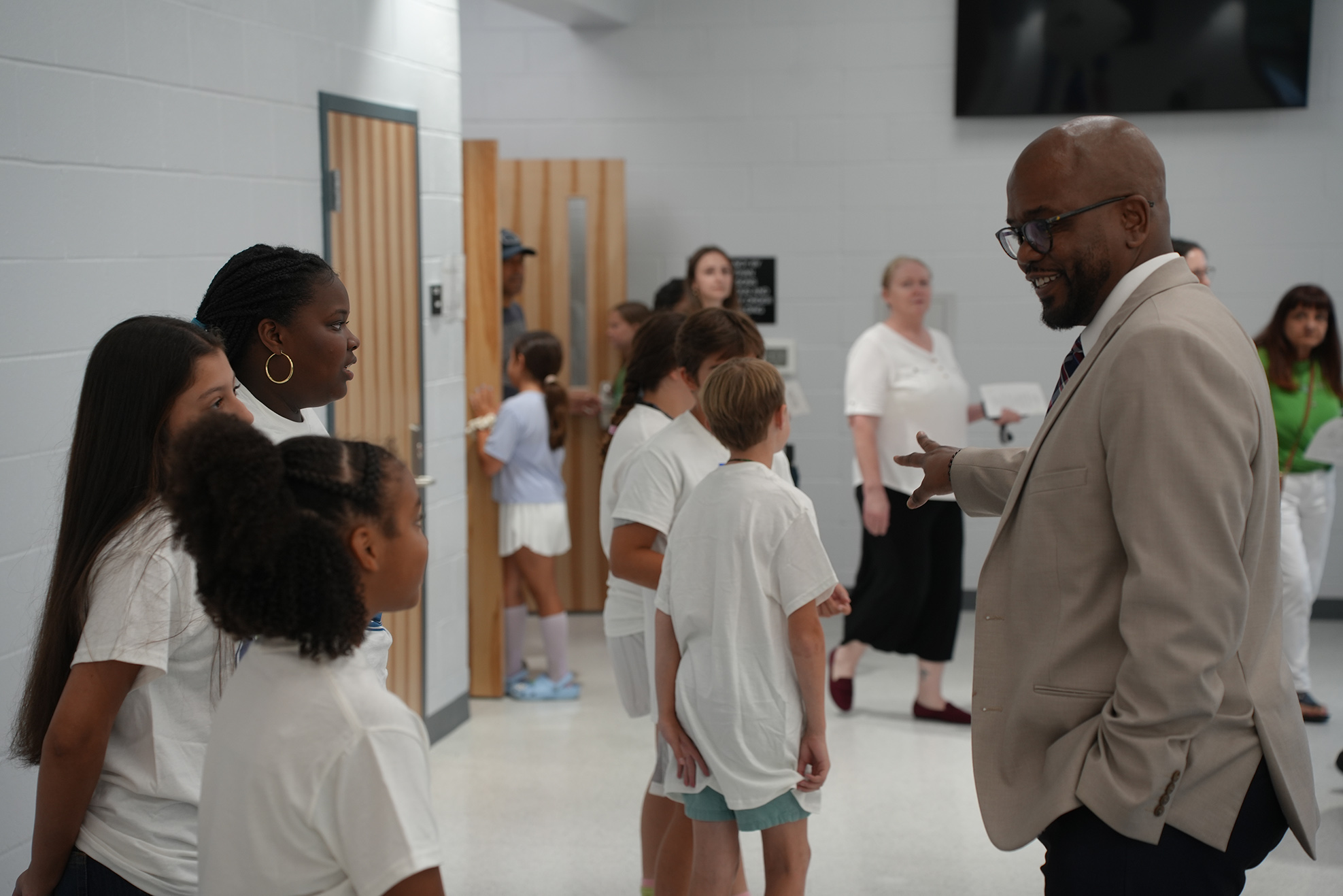 A man speaks to a group of children in a hallway; some children wear sneakers, and some wear socks.