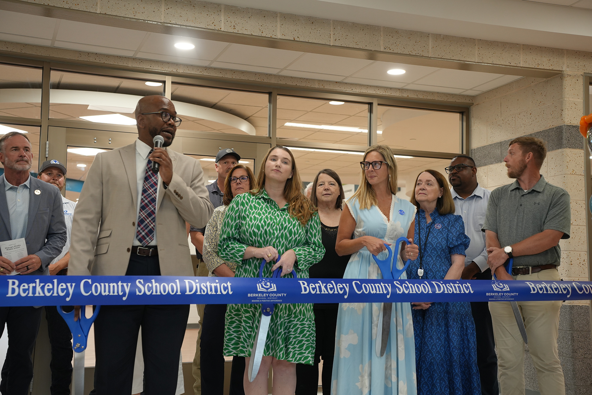 People standing by a ribbon. A man speaks into a microphone. Women hold scissors for cutting ribbon.