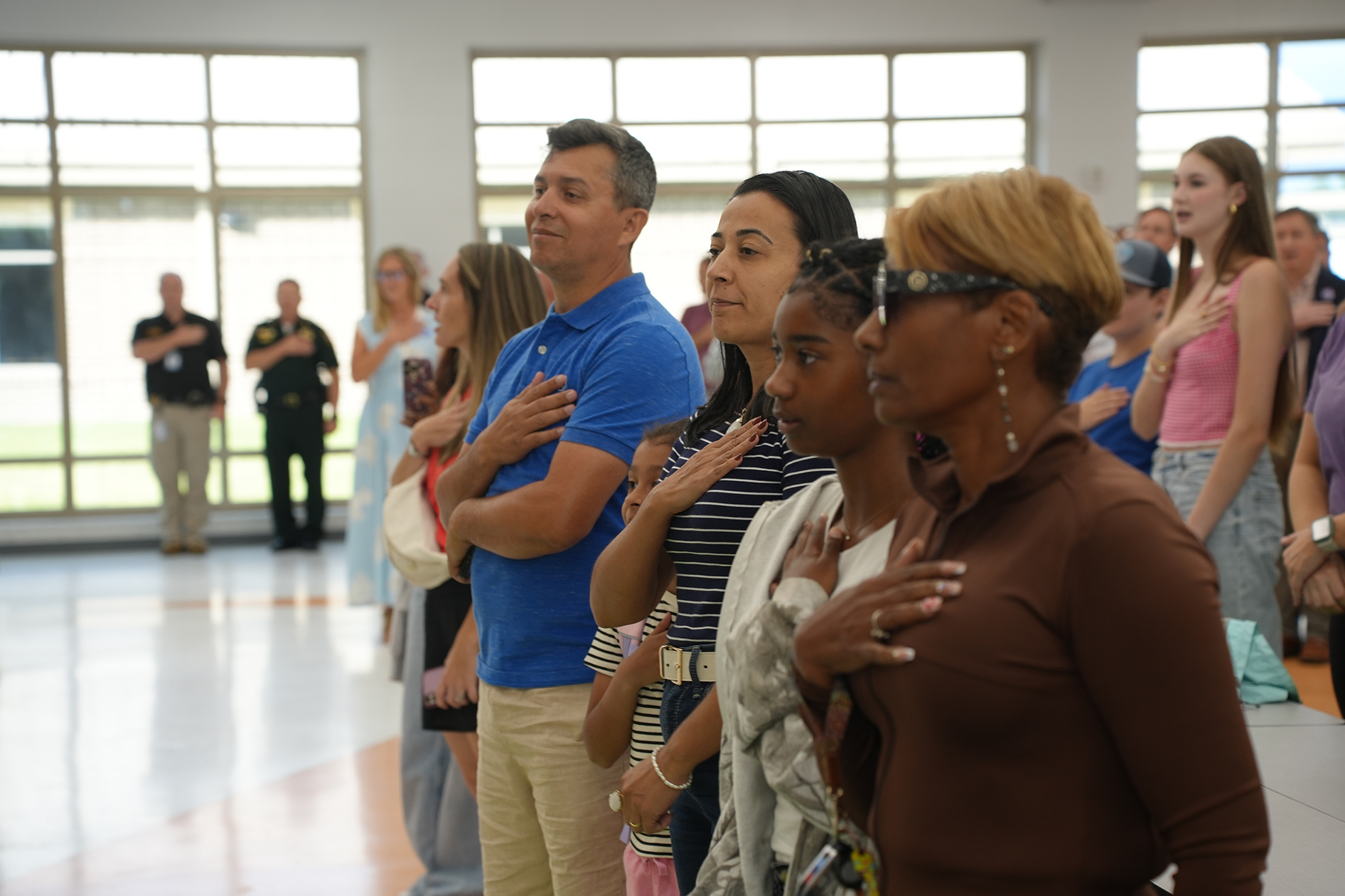A group of people stand in a room, some with hands over hearts. Two police officers stand in the background.