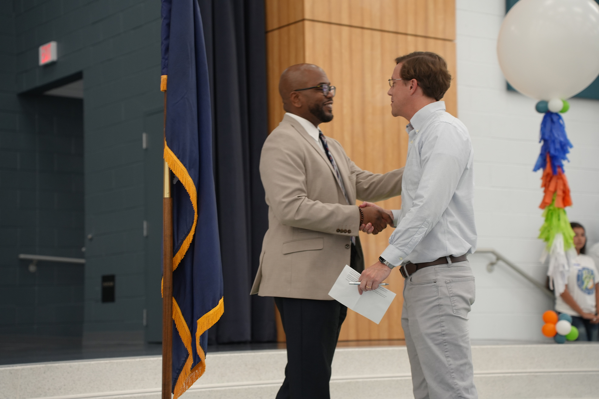 Two men shake hands in a hall. One holds a paper and wears a tie. A flag and balloons are in the background.