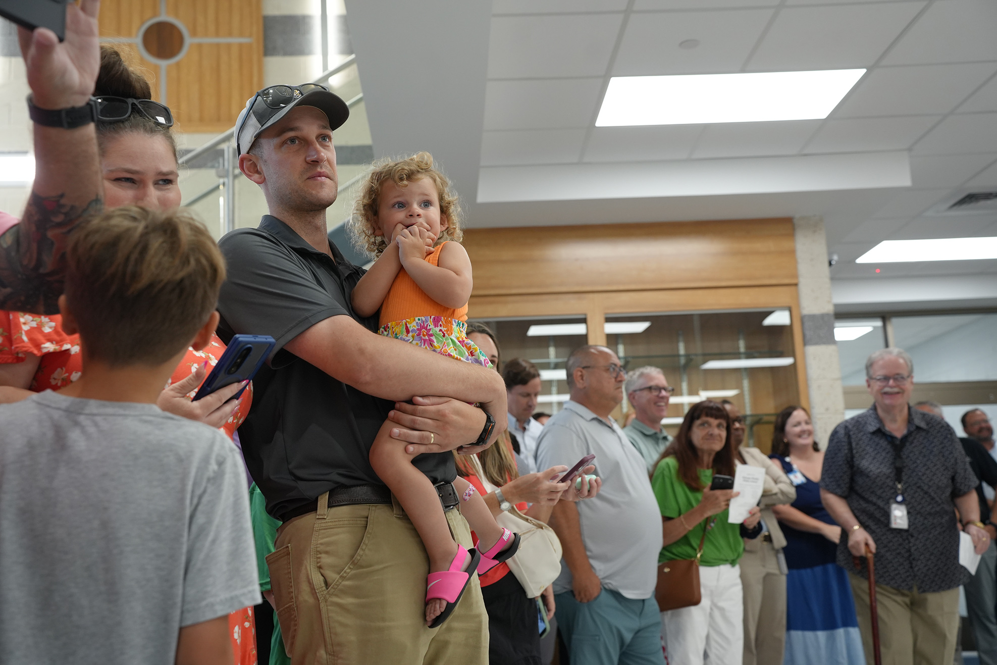 A crowd of people, including adults and children, stands indoors. A man holds a young girl while others look at phones.