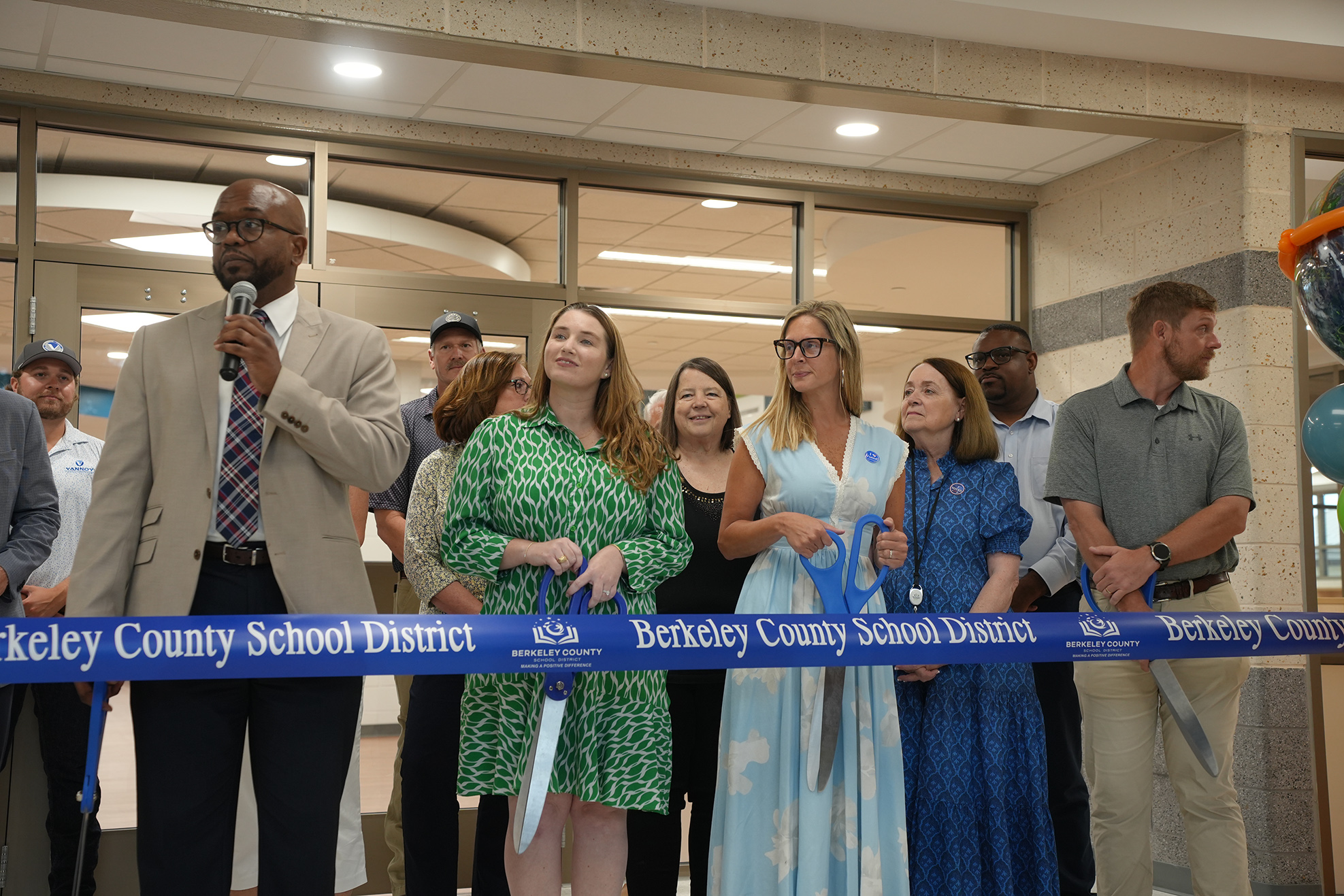 People stand around a blue ribbon. A man holds a microphone. A woman holds scissors and cuts the ribbon.