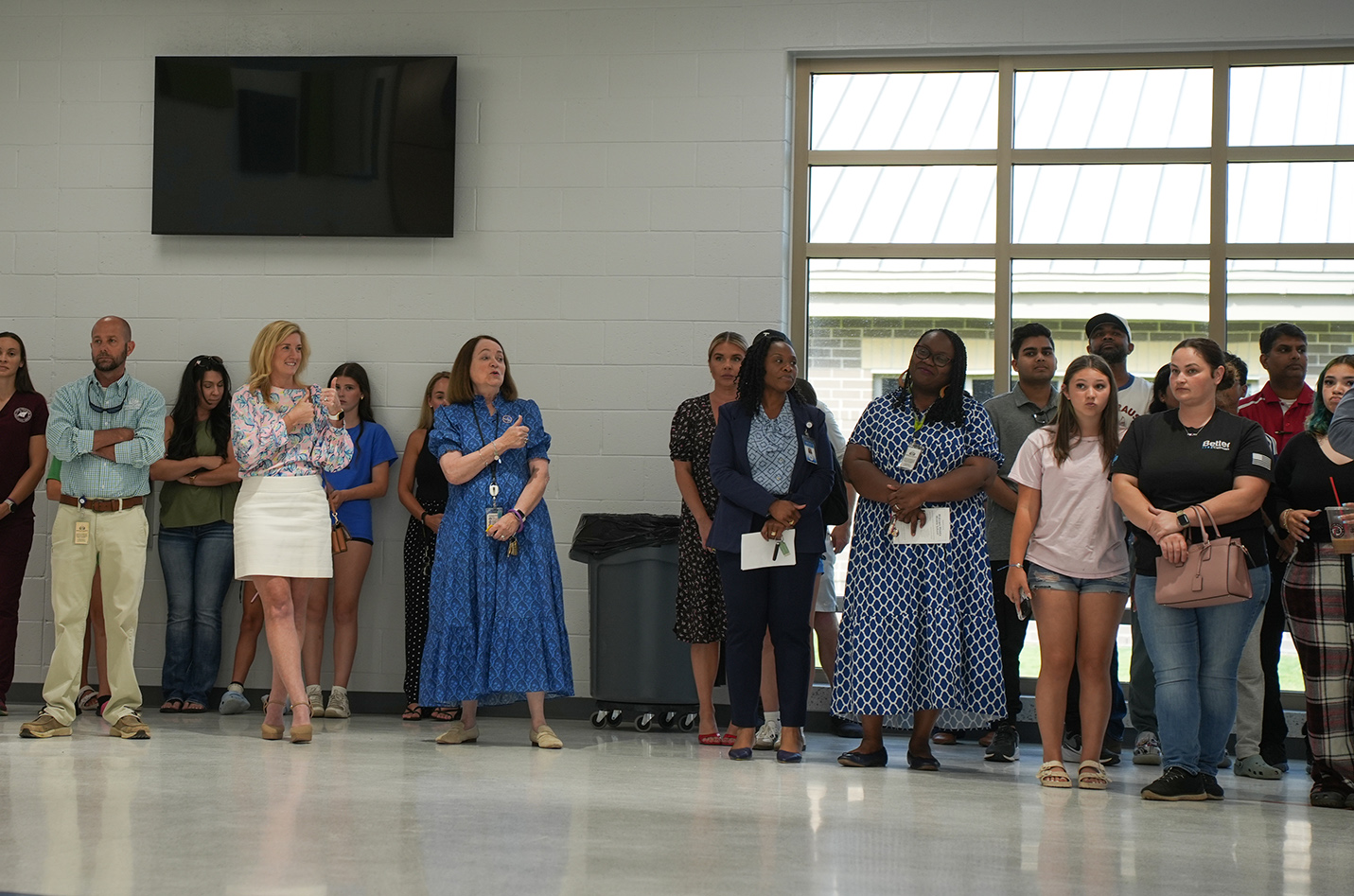 A group of people stand in a room, facing forward. Some hold papers. A trash can and monitor are on the wall.