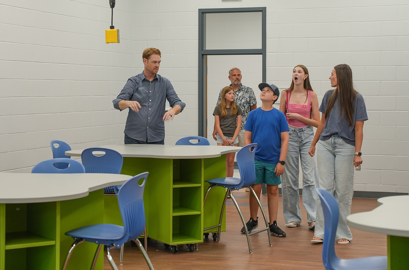 A group of people stands in a classroom with desks and chairs. A man speaks while a boy and others listen.