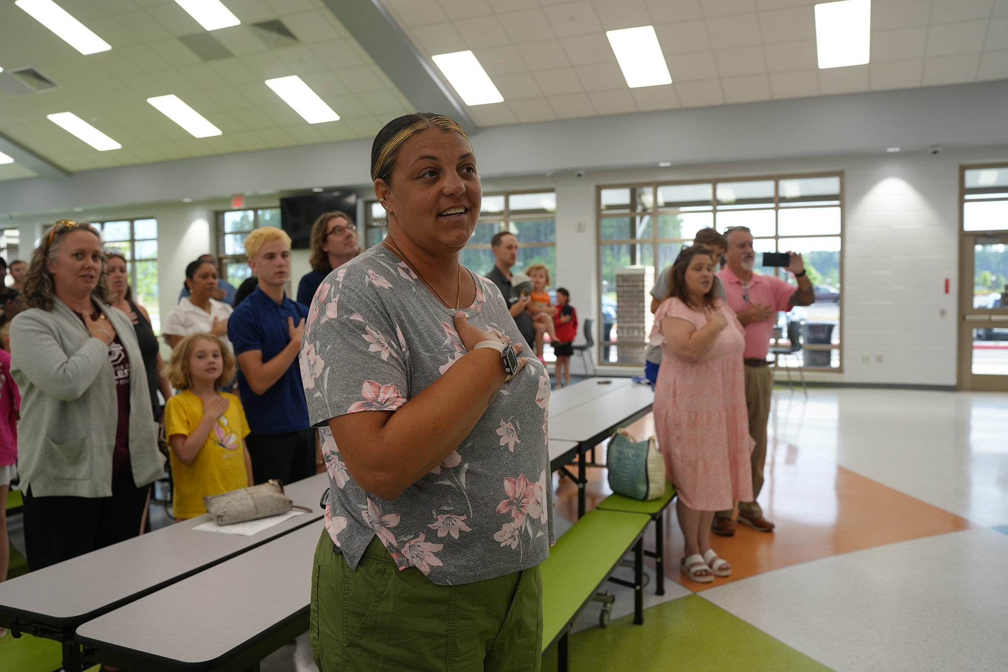 A woman wearing a floral shirt stands in a room with others, some with hands over hearts.
