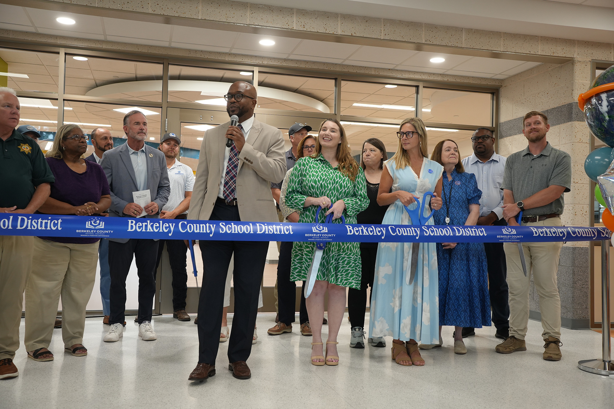 A ribbon-cutting event with people lined up. A man speaks into a microphone. Others hold the ribbon.