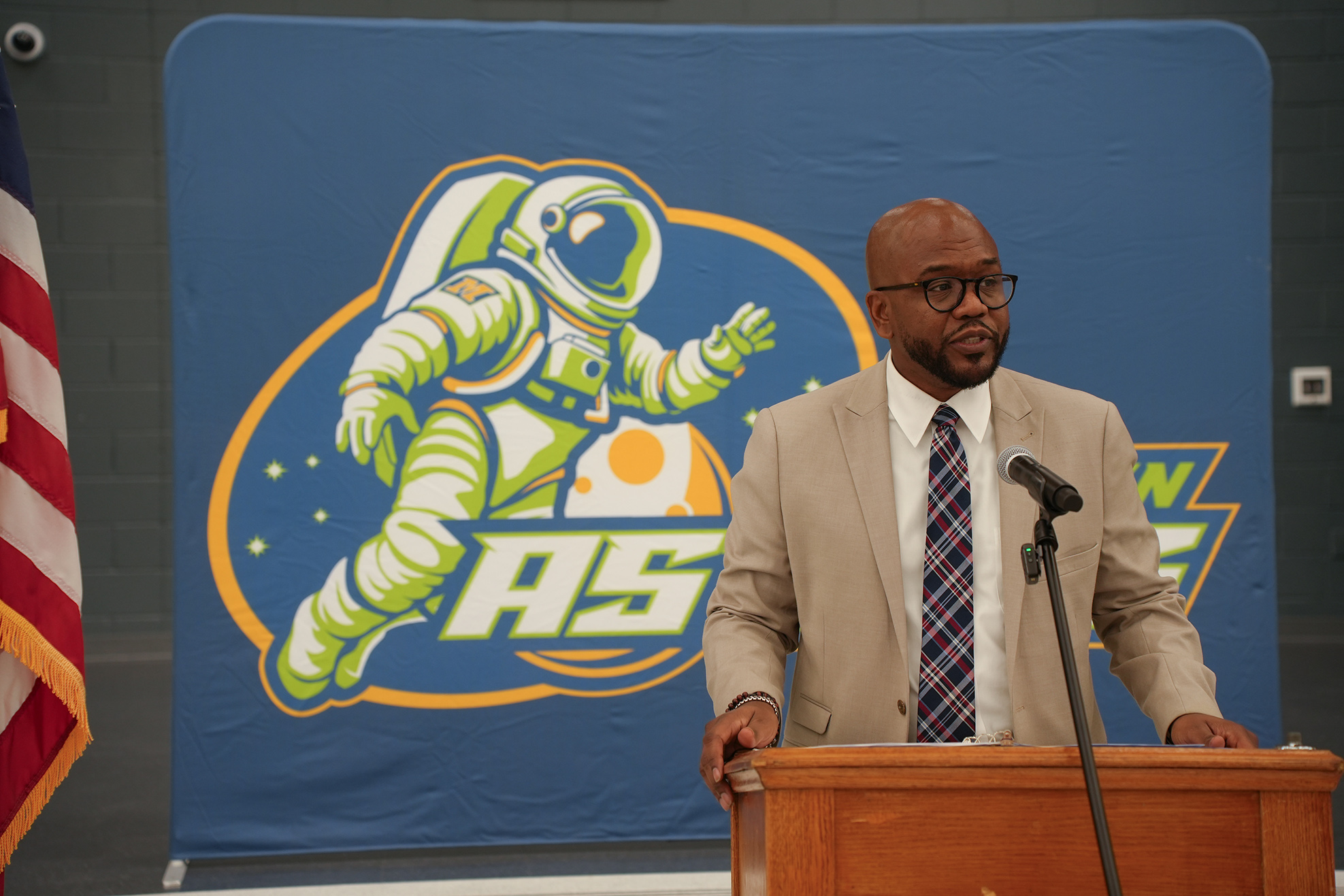 A man stands at a podium with a microphone, wearing glasses and a tie. Behind him, a large banner features an astronaut with text "AS" on a blue background.