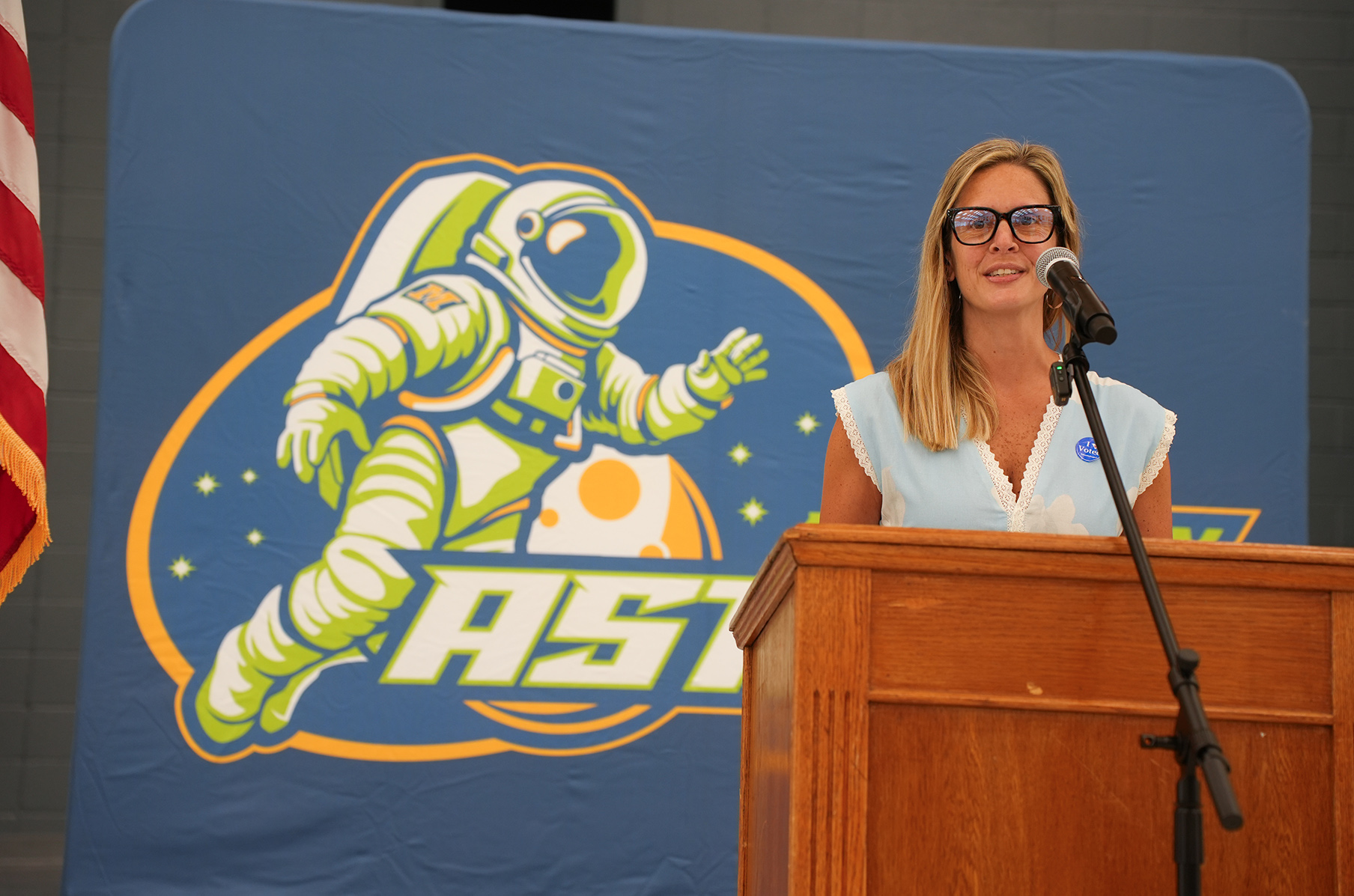 Woman standing at a podium with a microphone, an astronaut logo on a blue backdrop behind her.