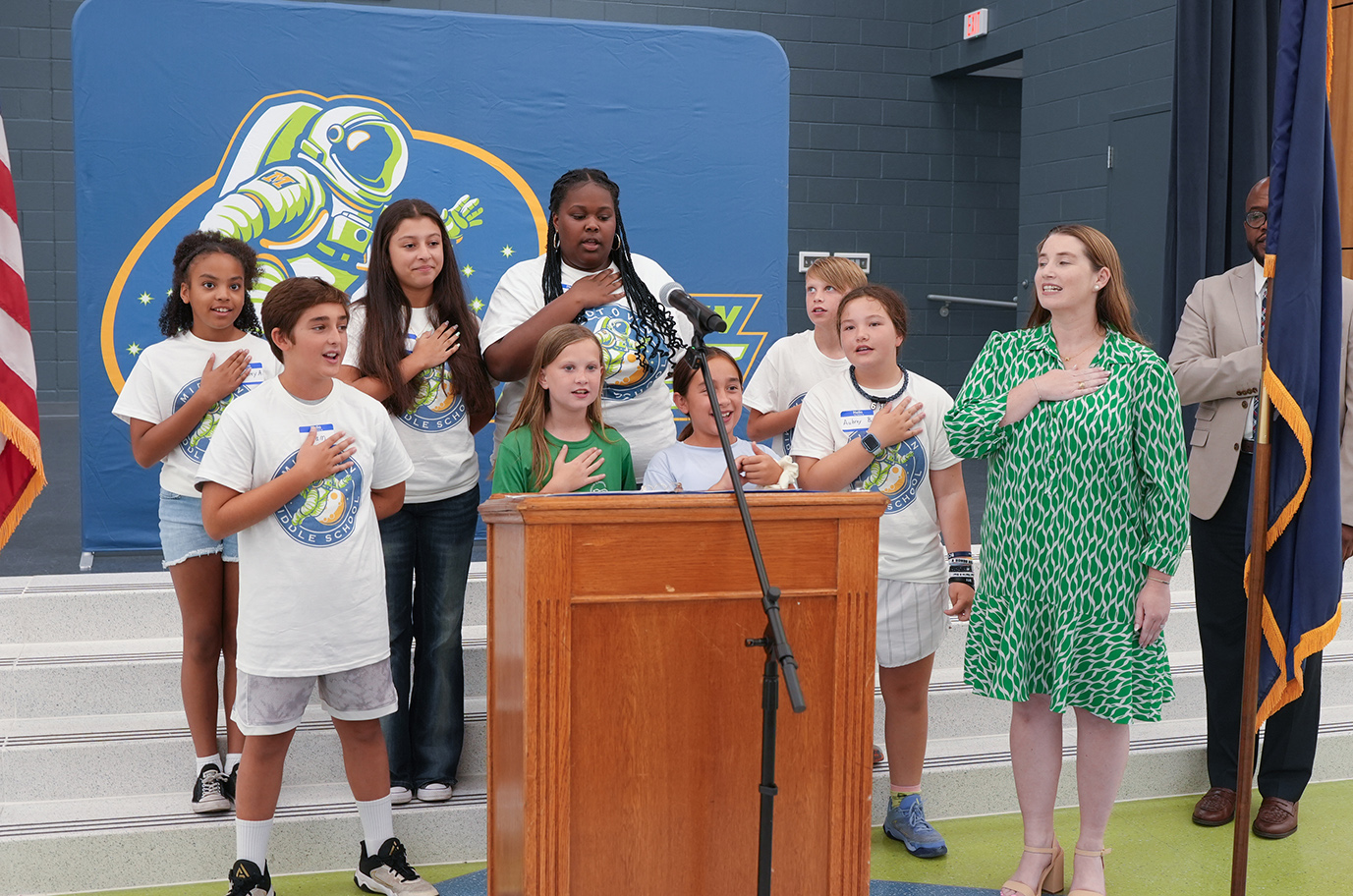 A group of children and adults stand on a stage, reciting the Pledge of Allegiance. A woman in green stands at the podium.