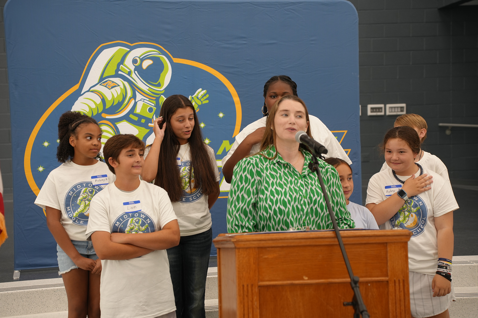 A woman speaks into a microphone at a podium while a group of young children stands behind her.