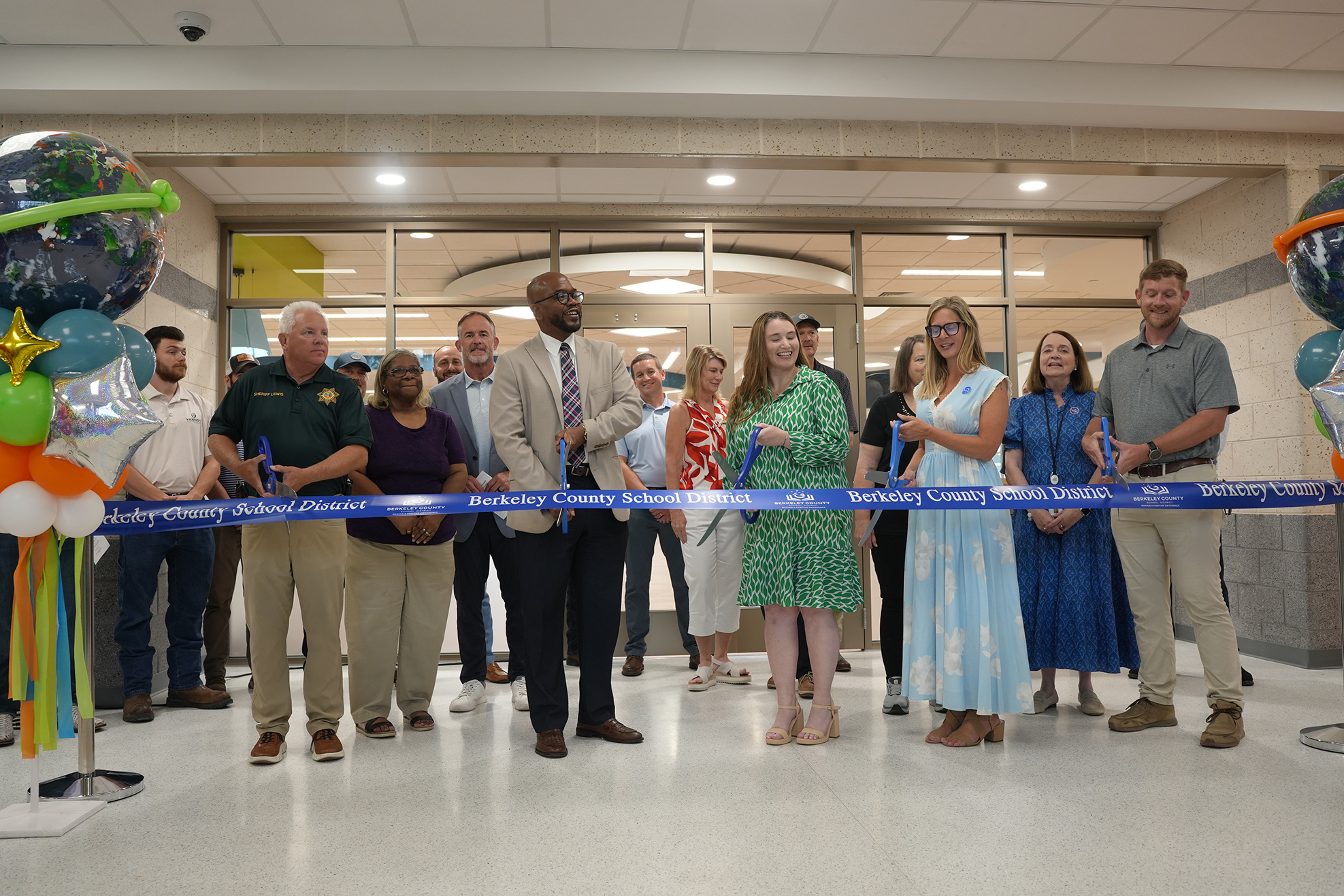Group of people standing around a blue ribbon, possibly attending an event. Some people are holding scissors.