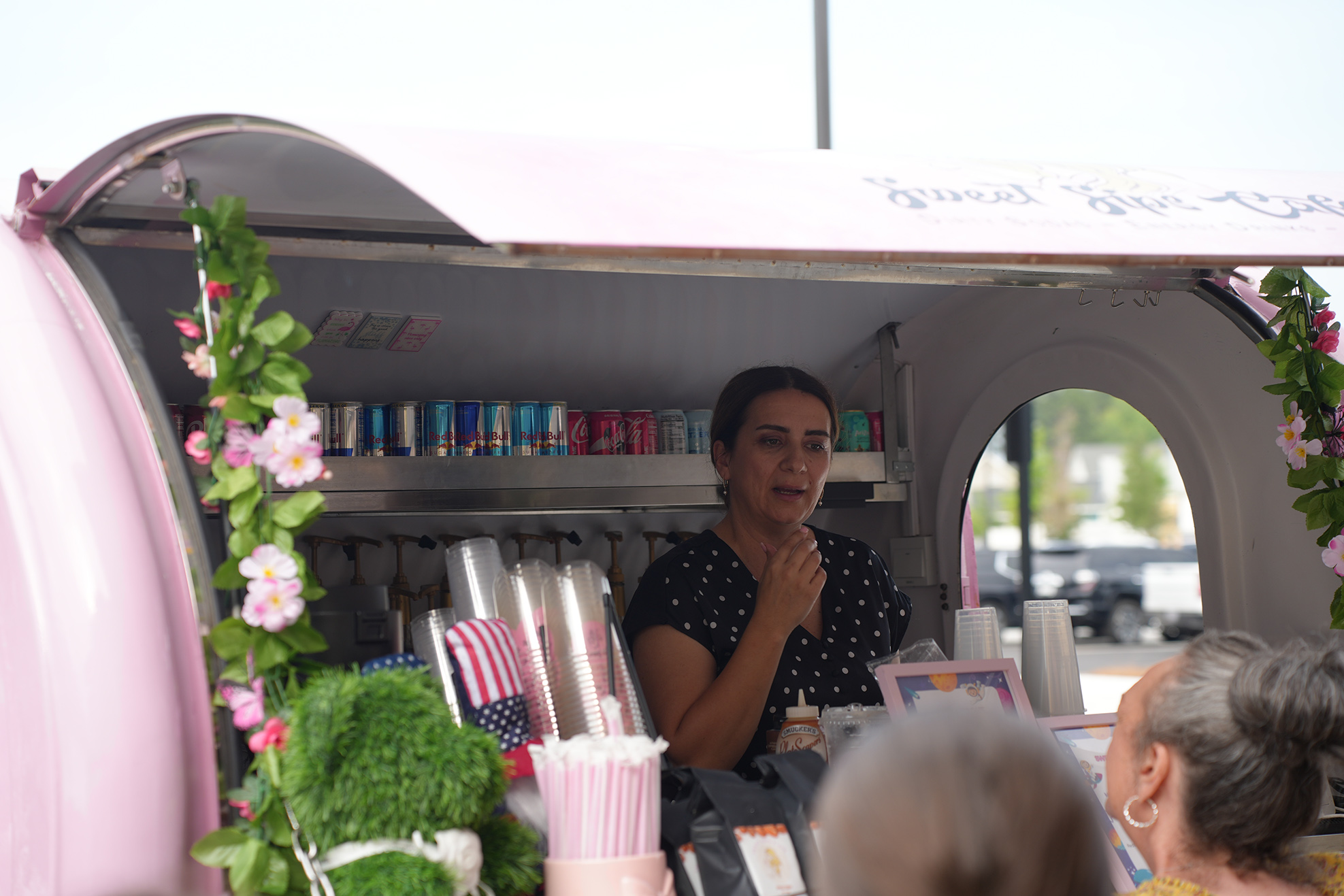 A woman stands inside a pink food truck with floral decorations, a shelf with drinks, and a US flag.
