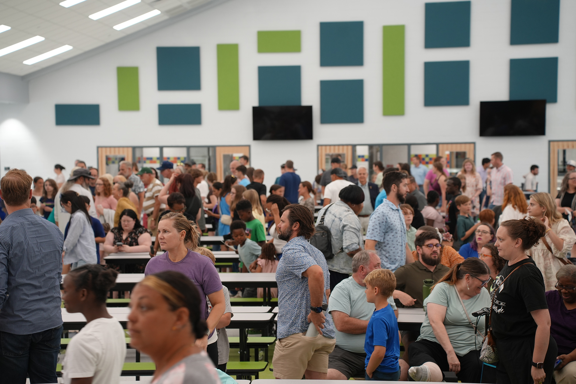 Many people sit at tables in a cafeteria with green-and-blue panels on the wall and two monitors.