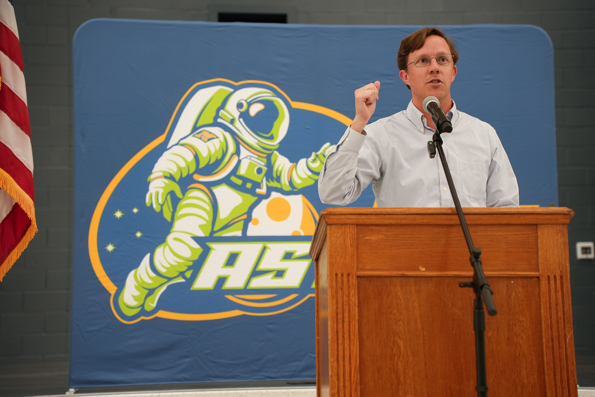 A man speaks at a podium with a microphone. A blue backdrop features a cartoon astronaut.