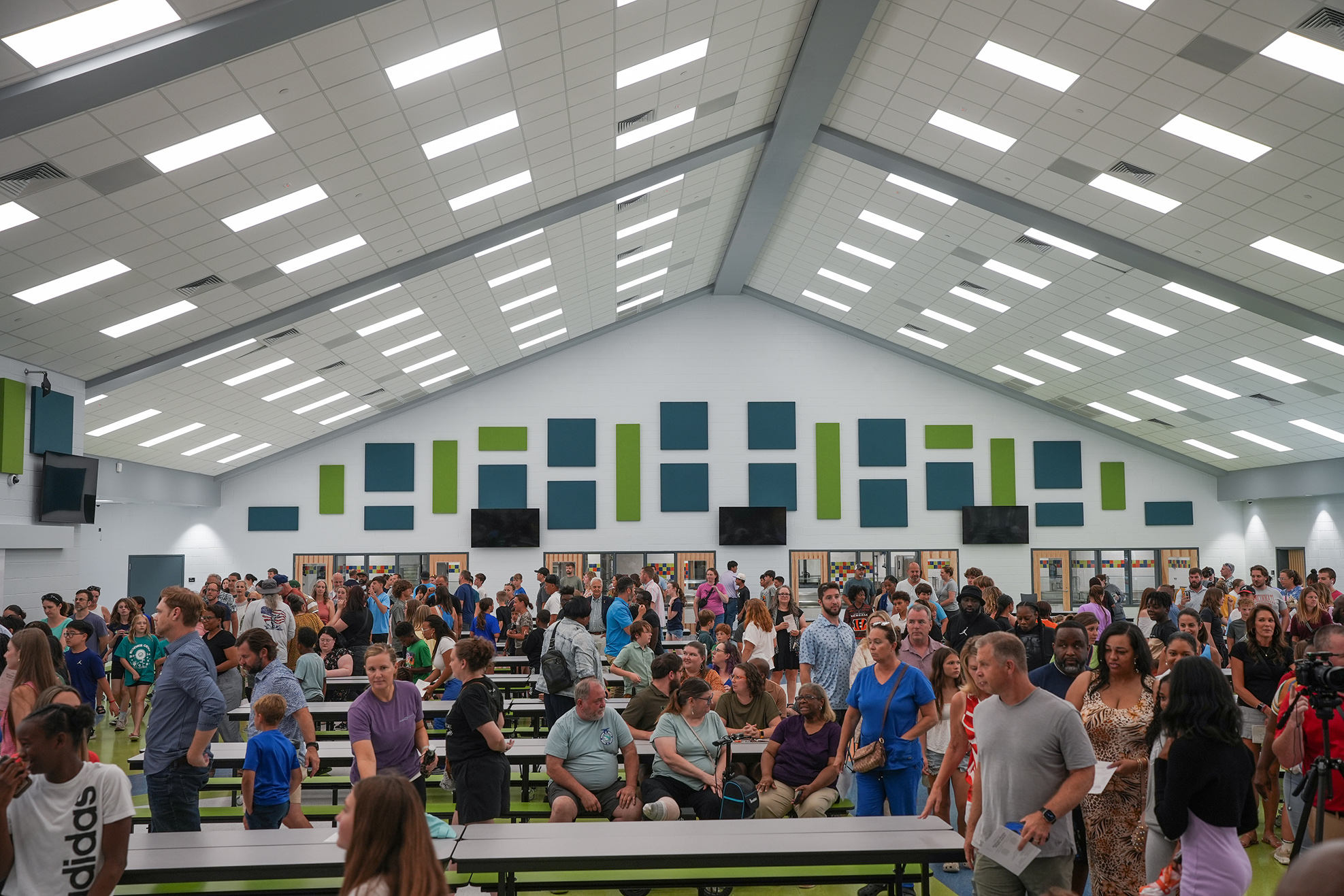 A large group of people in a cafeteria. Long tables are filled with people, and the room is well lit.
