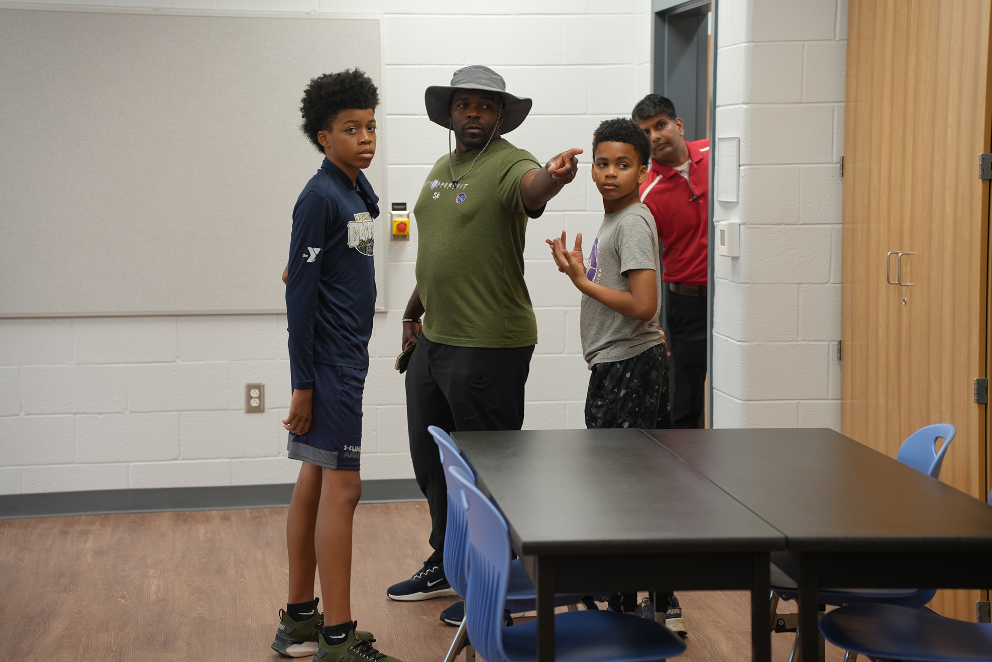 Four boys, one in a hat, stand in a classroom with a table. The room has wooden floors and white walls.