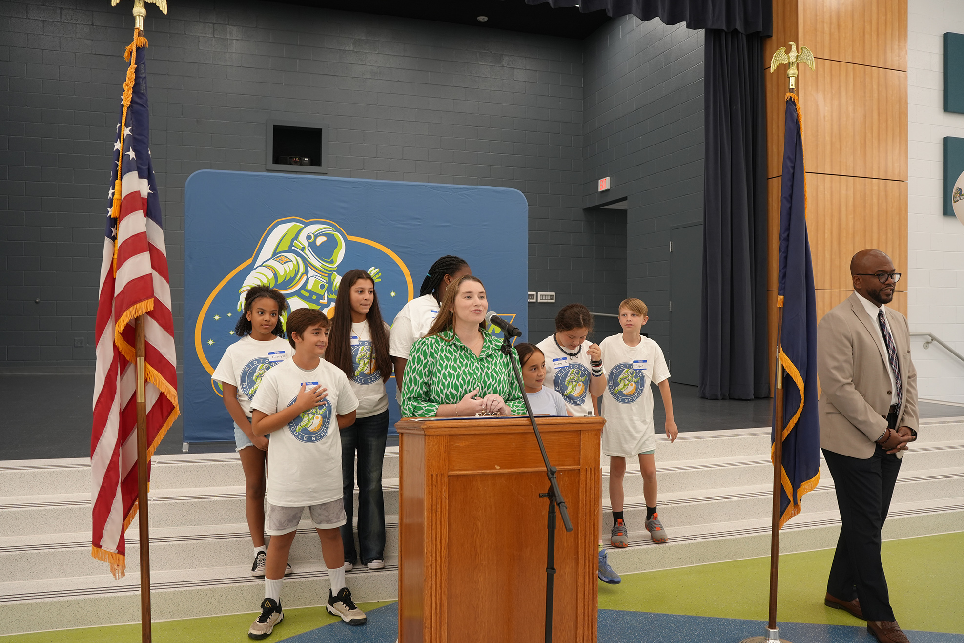 A woman speaks at a podium while standing before a group of children in white shirts. Two flags stand on either side.