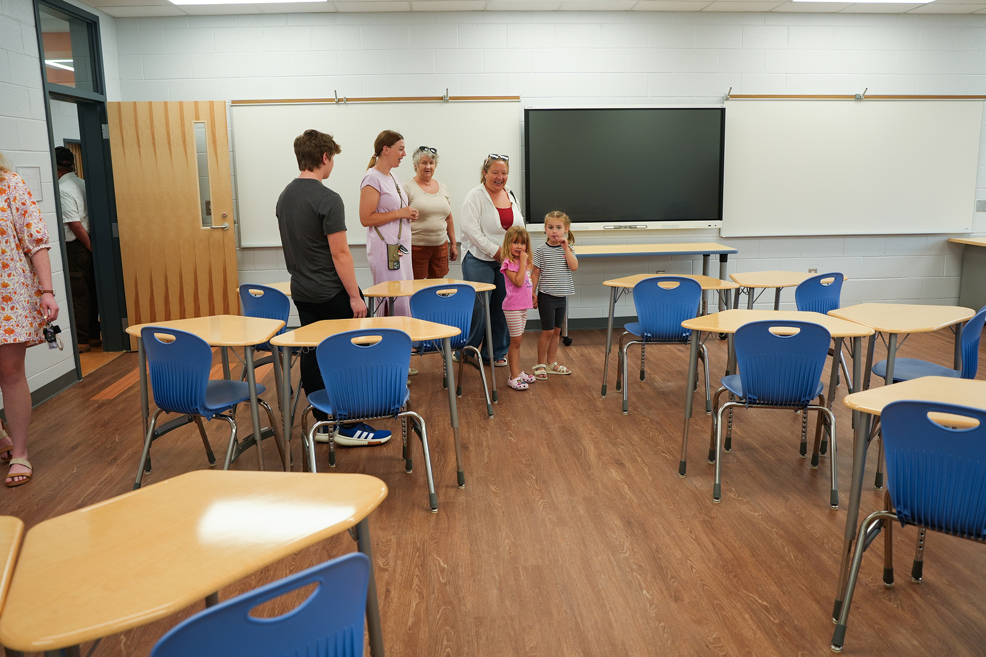 People in a classroom with wooden floors, blue chairs, and tables, a monitor on the wall, and a door.