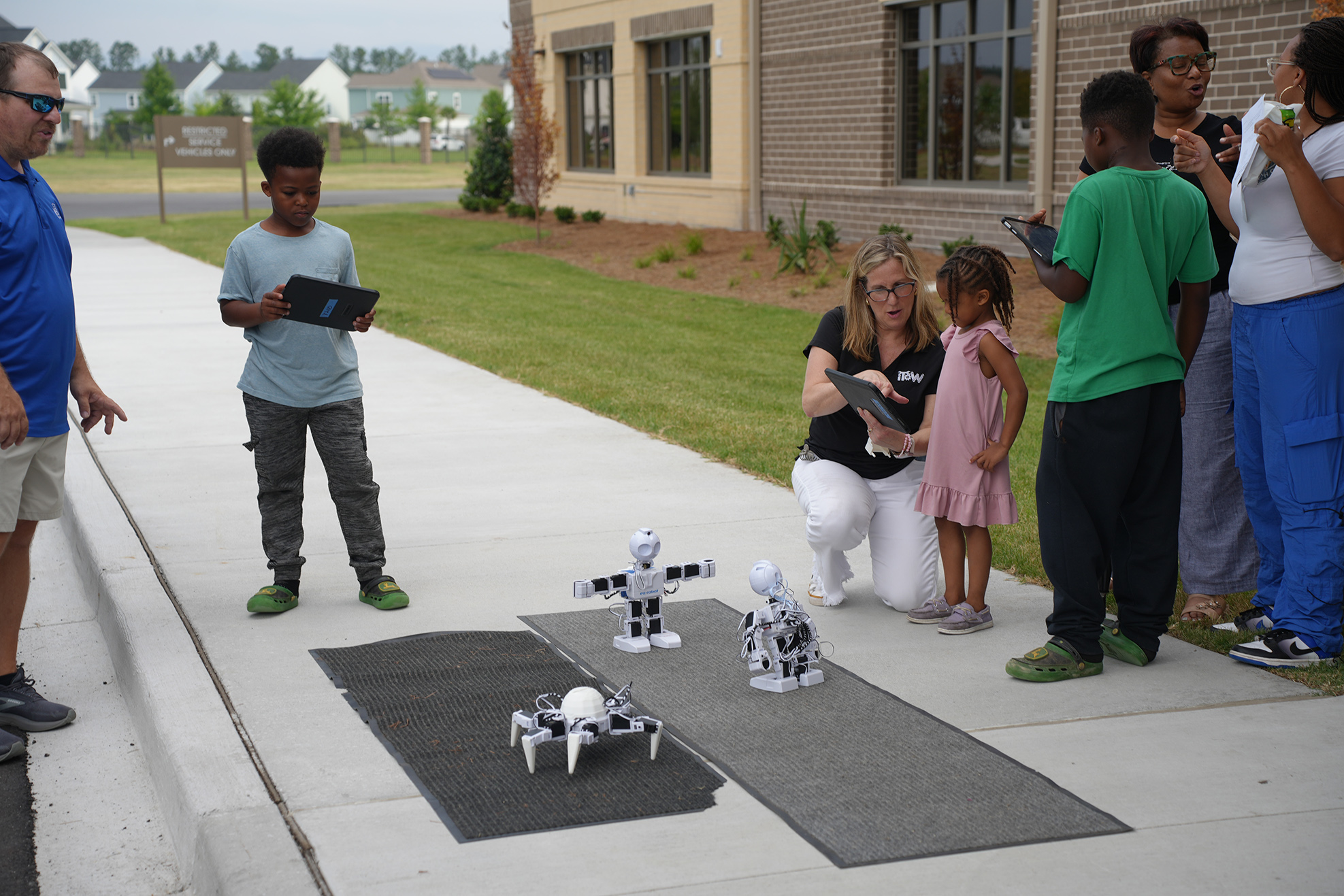 People in front of a building watch a demonstration of small robots on a sidewalk.