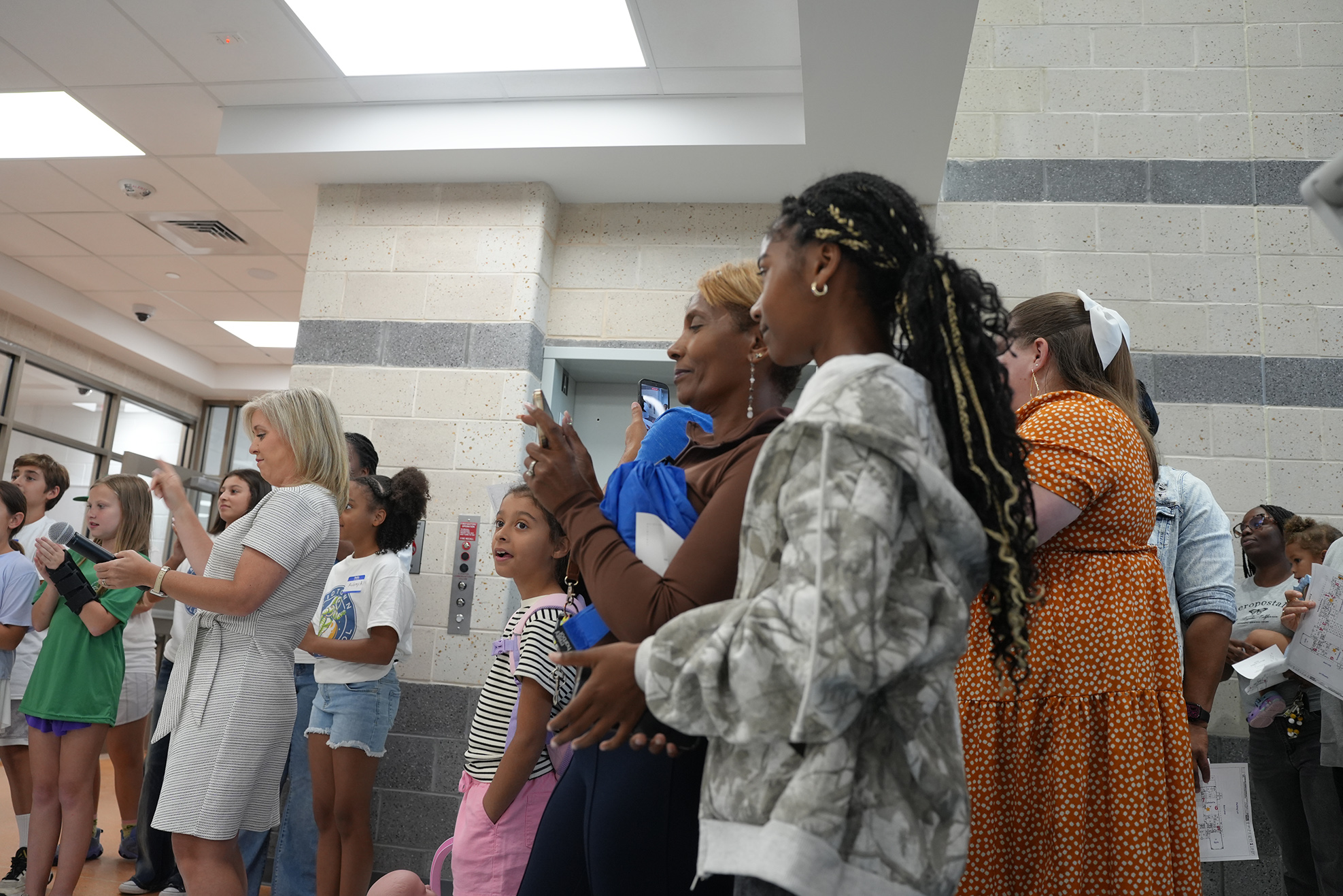 A group of people stands indoors. Some are looking upward. Some wear casual clothing. A person holds a blue cloth.