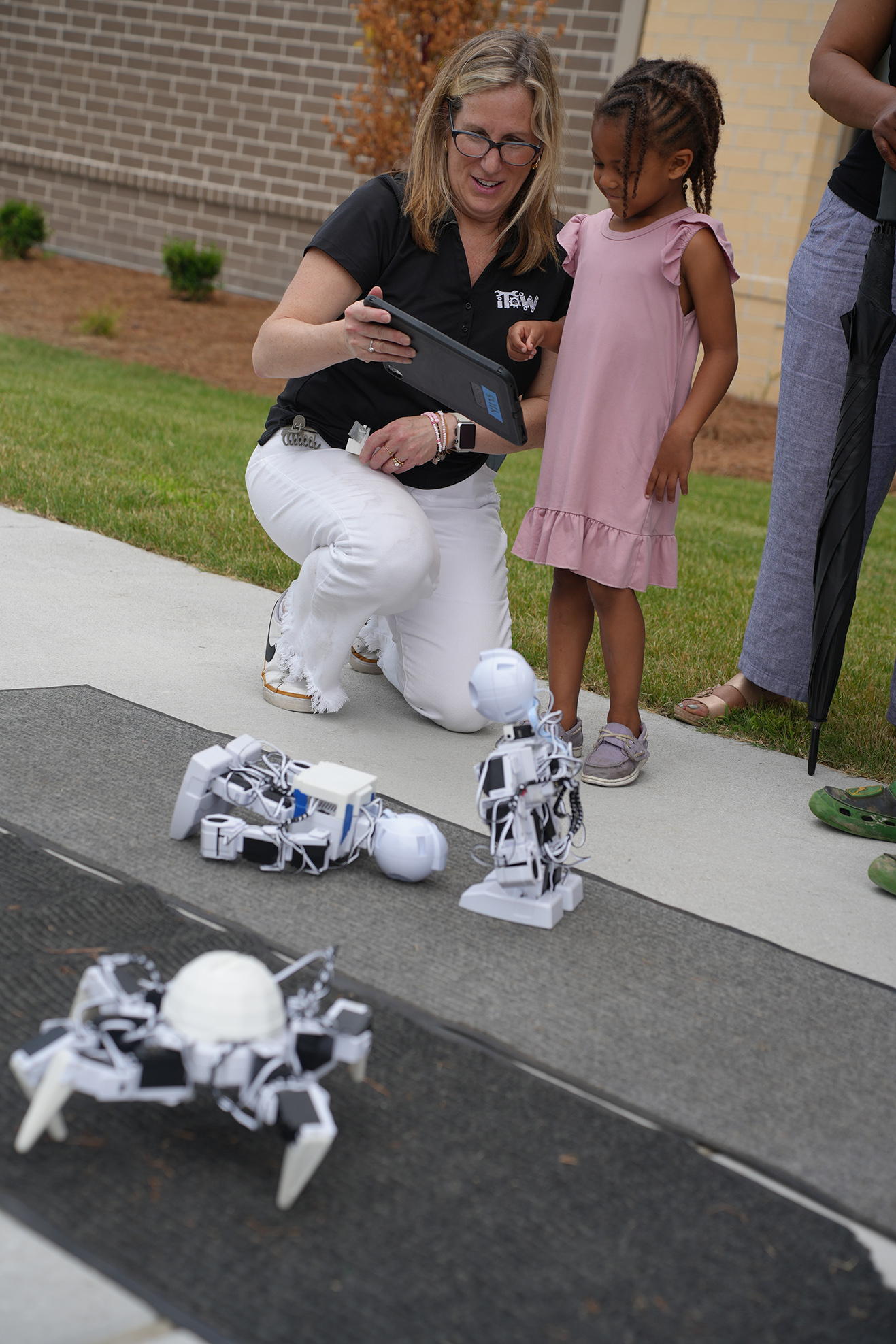 A woman in a black shirt crouches with a tablet, and a girl in pink looks at robots on the sidewalk.