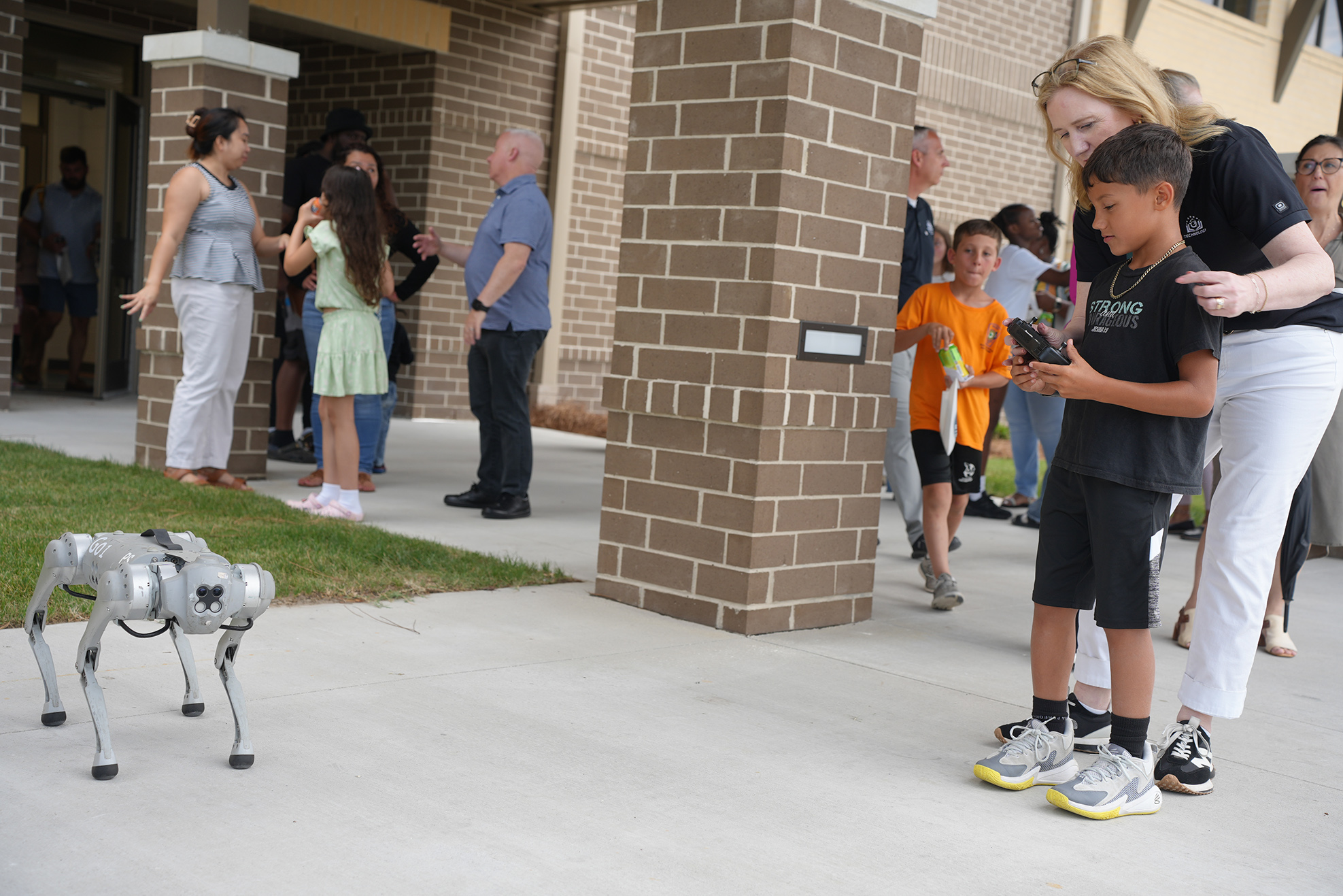 A woman and child interact with a robotic dog while others gather outside a brick building.