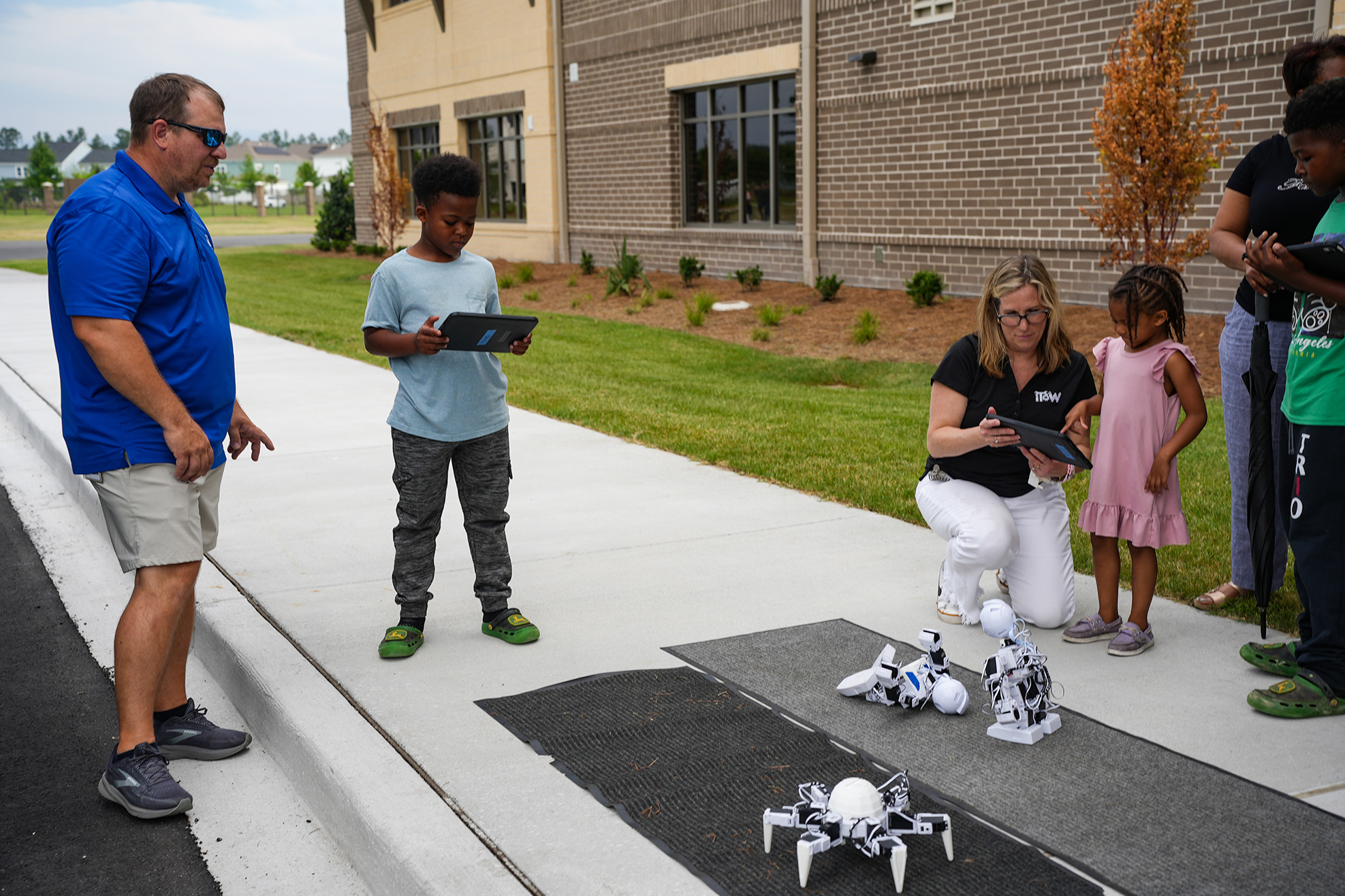 A group of people on a sidewalk near a building. A woman and a child are looking at robots.