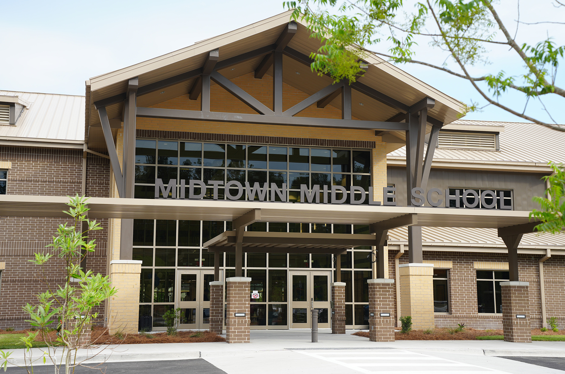 A school building with a metal roof. Glass windows and doors. Large sign reads "MIDTOWN MIDDLE SCHOOL". Trees and plants outside.