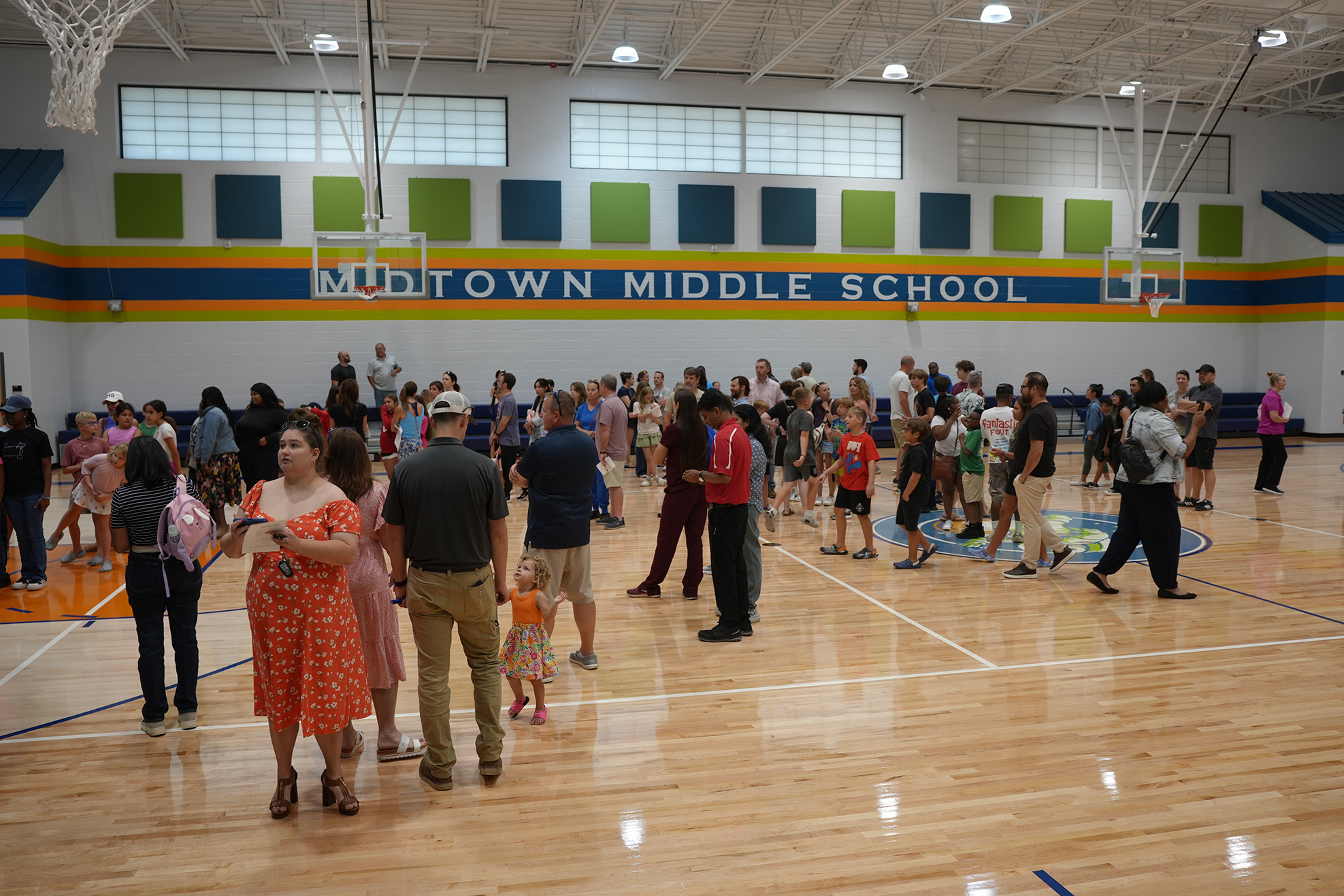 A gym filled with people. A blue, orange, and green wall with "Midtown Middle School" written on it.