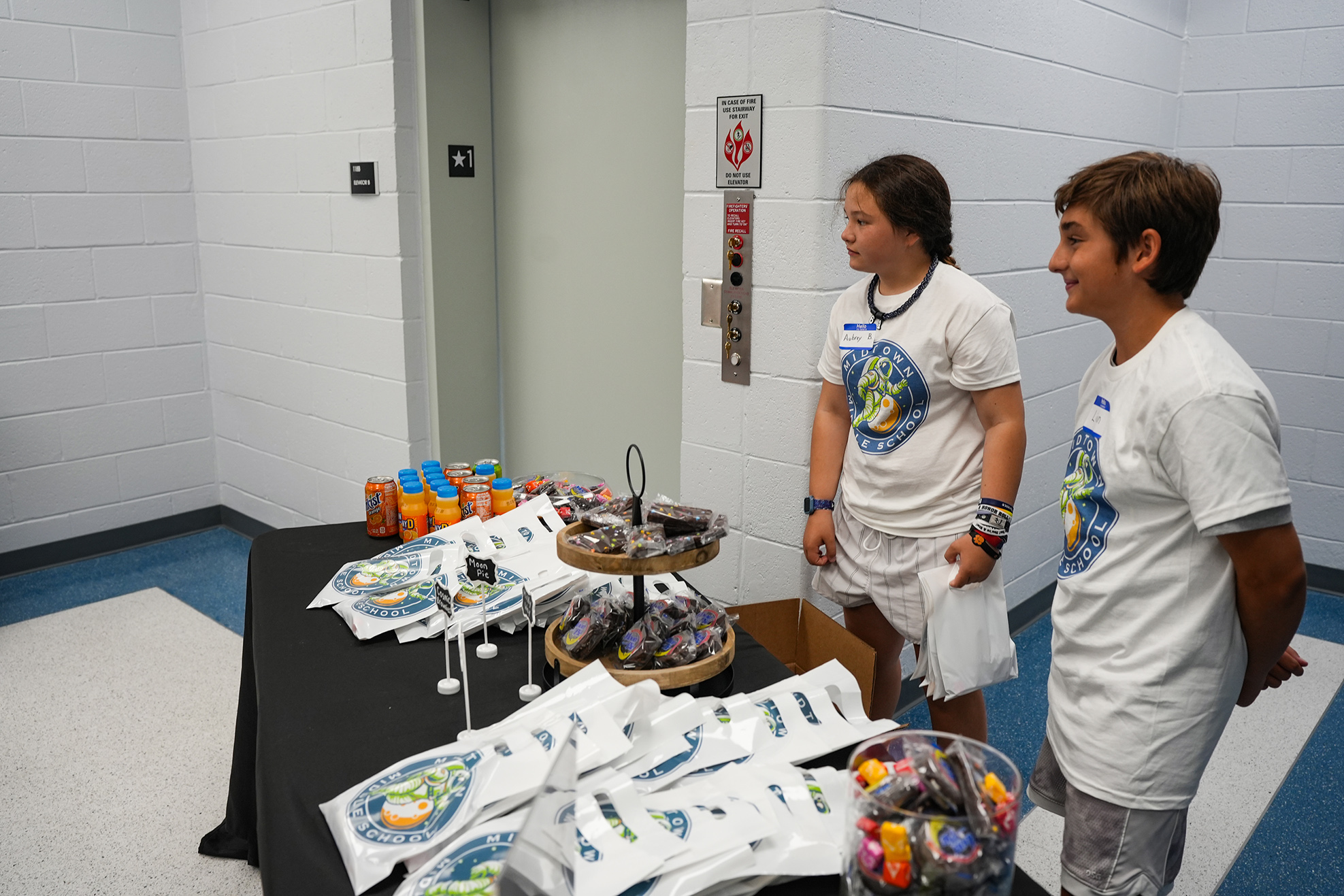 Two people in white shirts stand by a table with merchandise, a bowl of candy, and a white box.