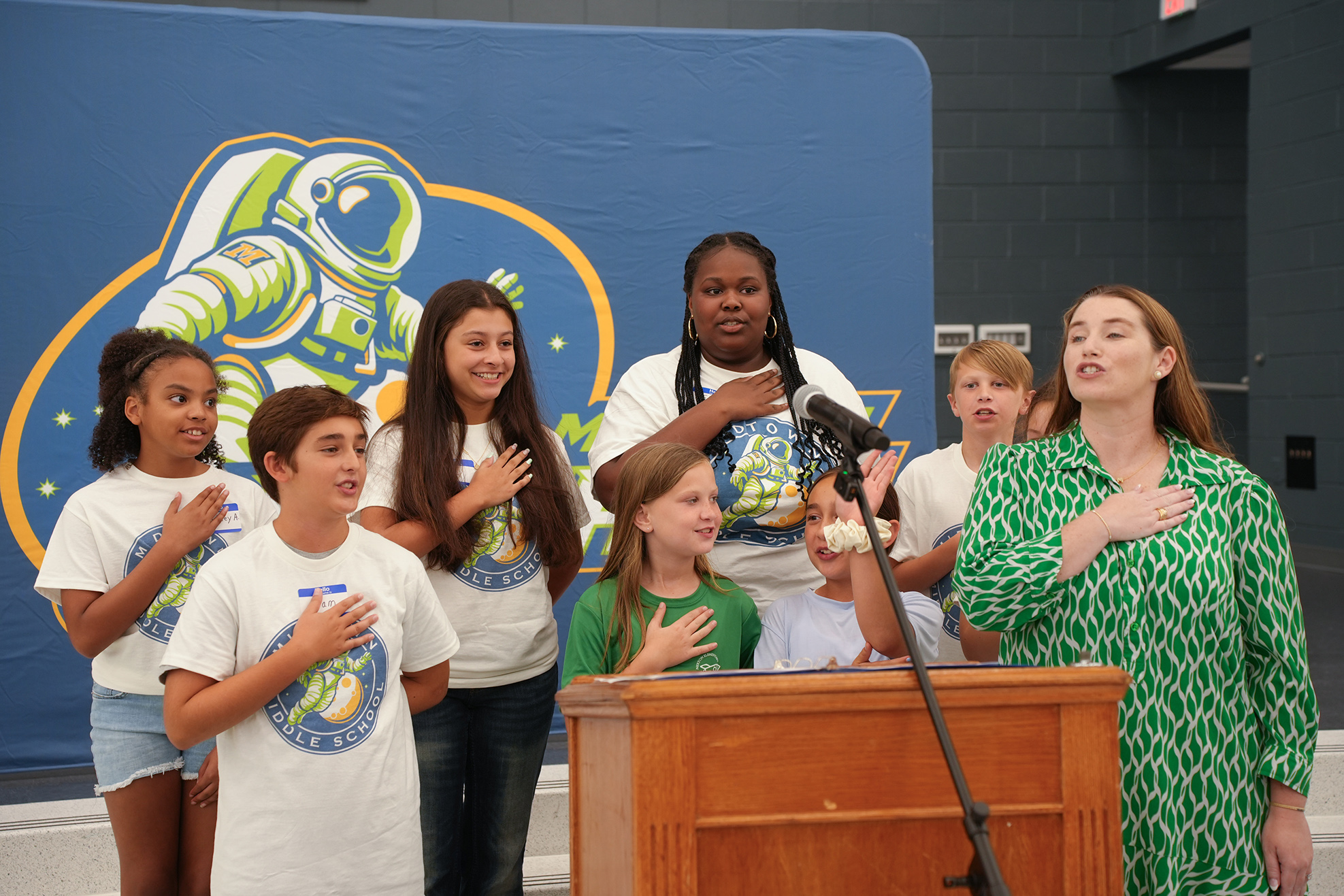 Group of children and adults in white and green clothes standing by a podium, with a microphone, and a blue backdrop.