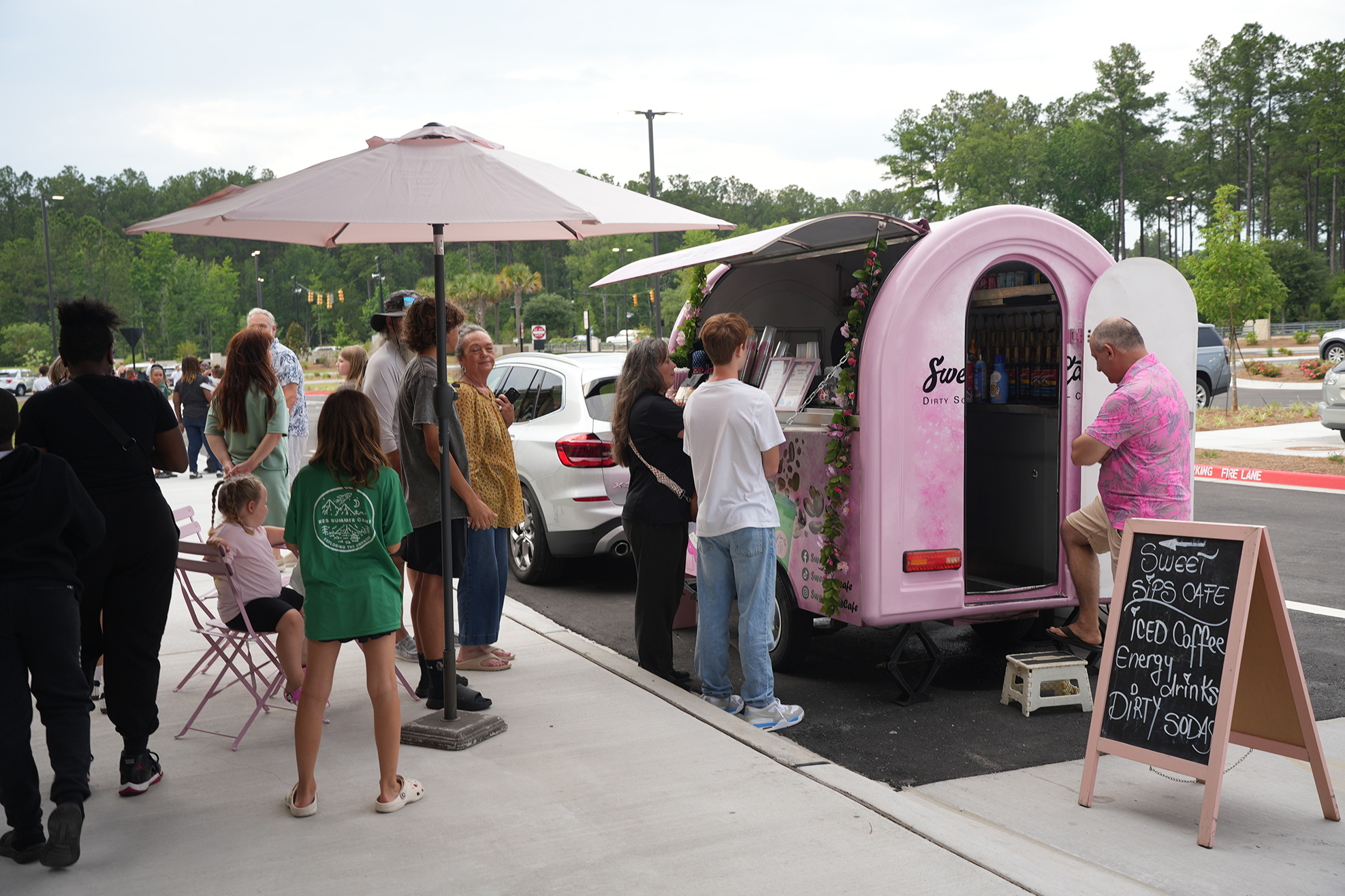 People standing in line at a pink food truck with pink flowers on it. A menu board is beside the truck.