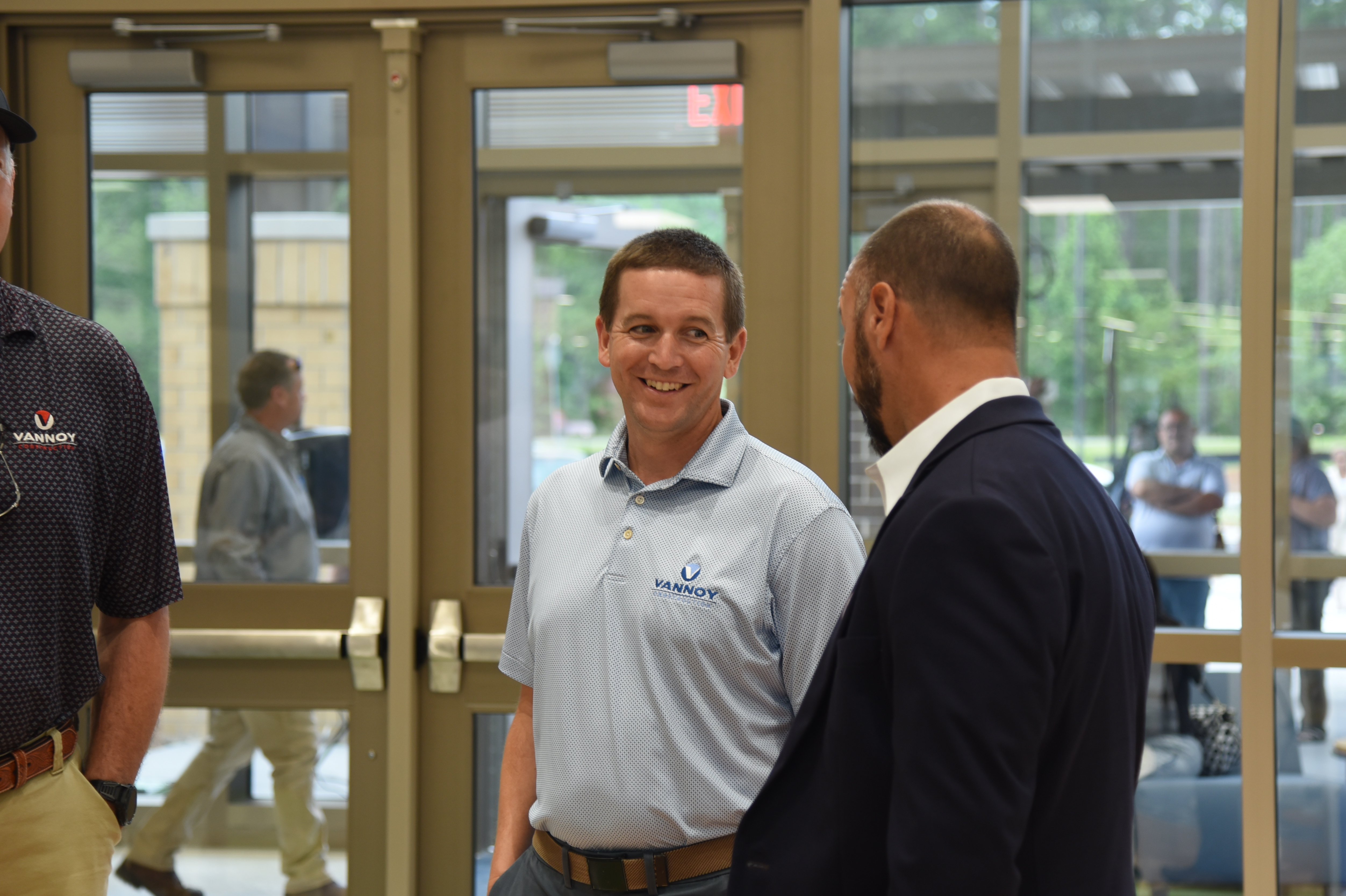 Three men in a corridor; one smiling and two in business attire conversing. Behind them, a man walks through a glass door.