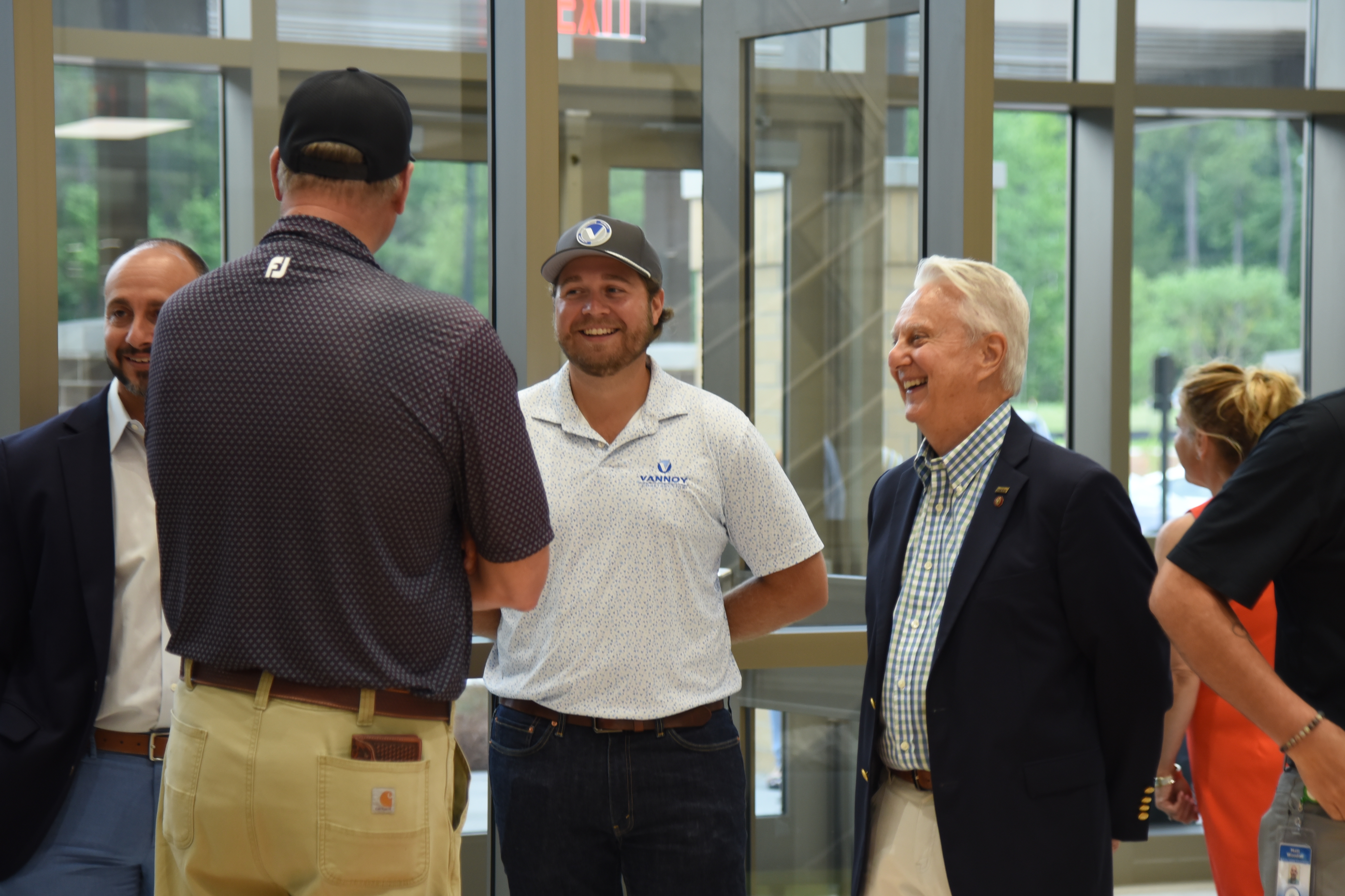 A group of people stand in a lobby. One wears a black cap and brown pants, another a white shirt and beige pants.