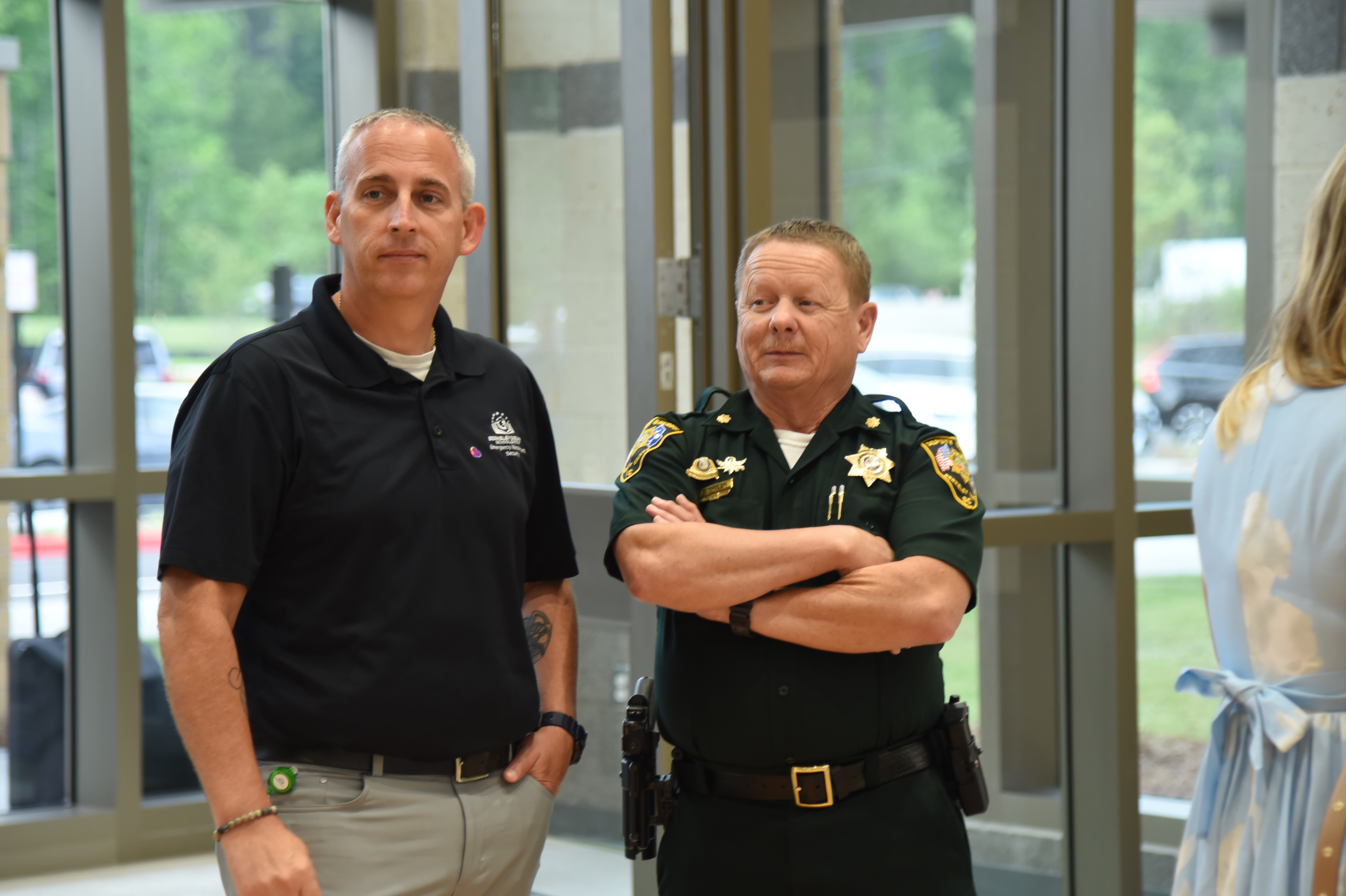 Two men in uniforms stand in front of a building; one has crossed arms and the other has a belt.