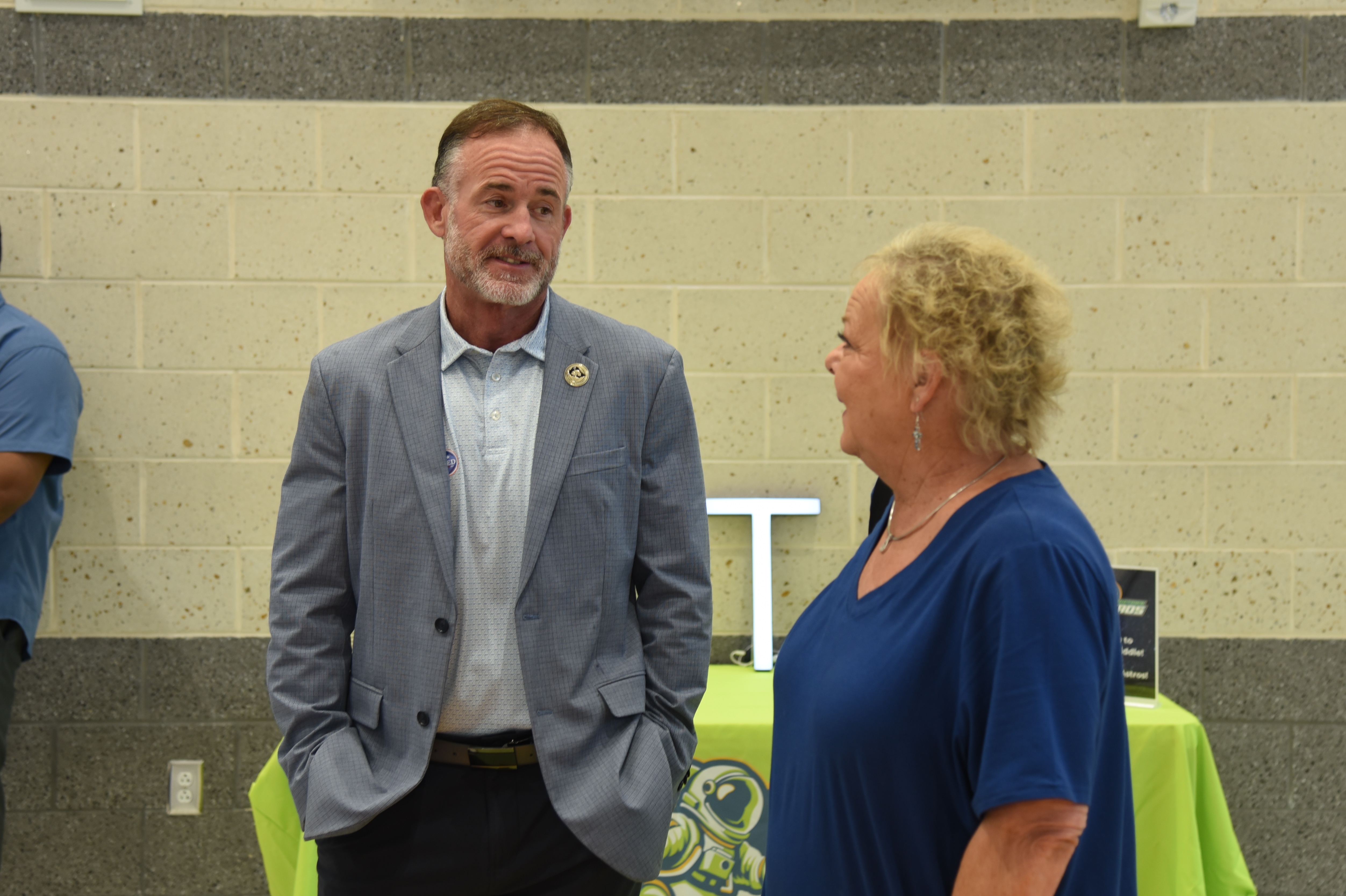 An adult man in a gray suit speaks with a woman in a blue shirt in a room.