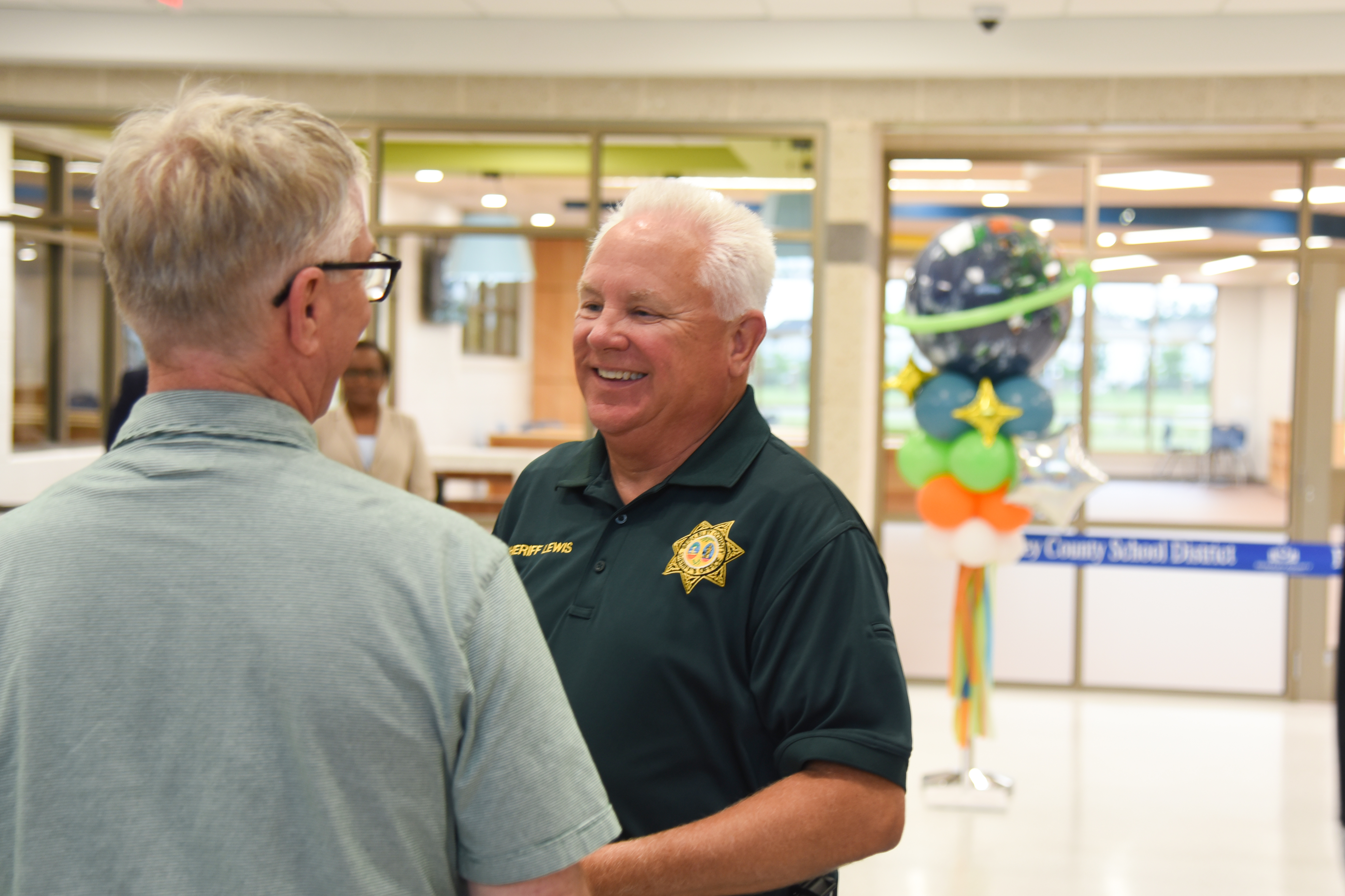 Two men converse inside an office. One wears a green polo with a logo. Behind them, a colorful balloon centerpiece.