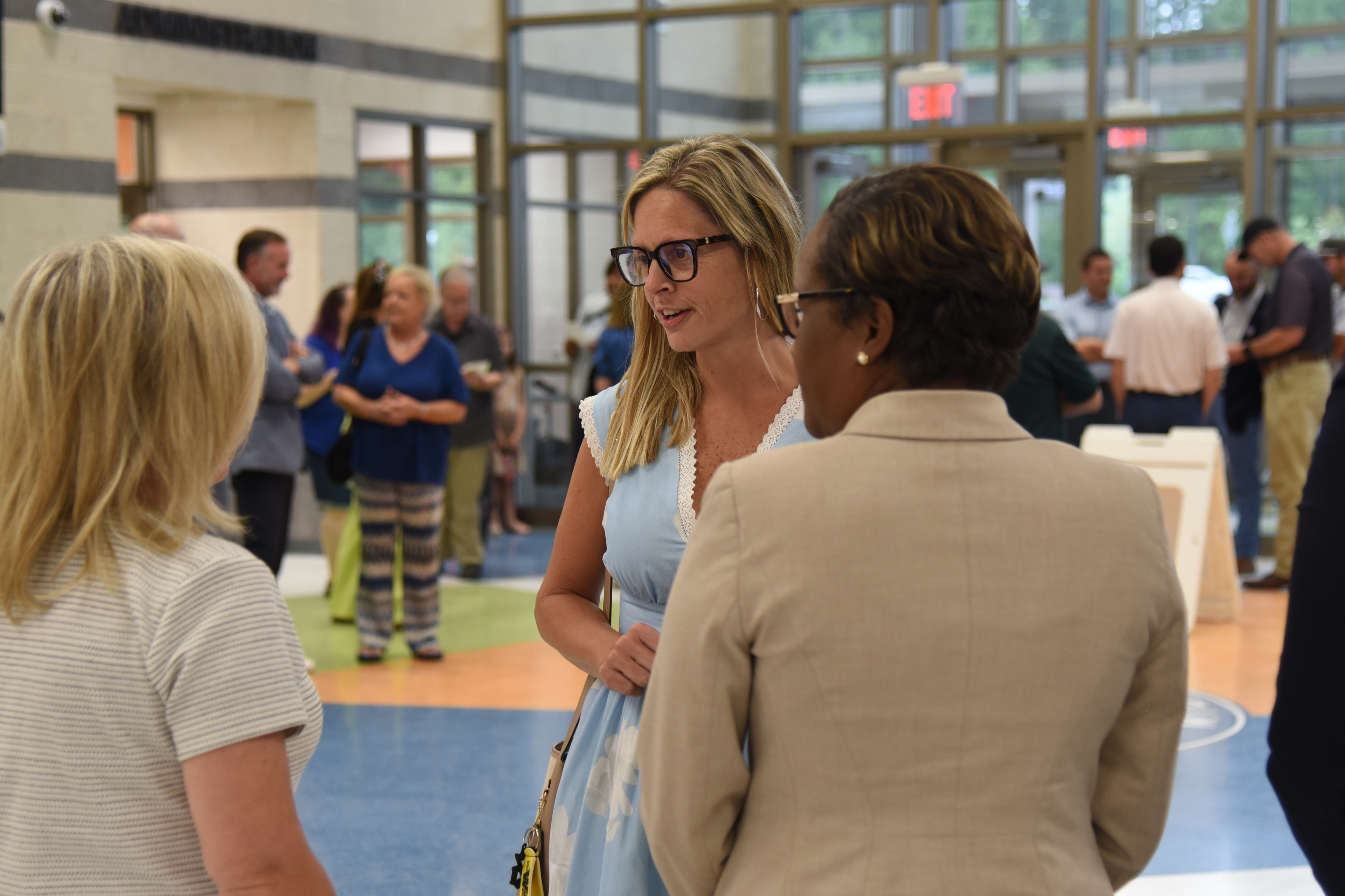 Several adults converse in an indoor area with blue flooring and glass walls. Some people in the background stand near white tables.
