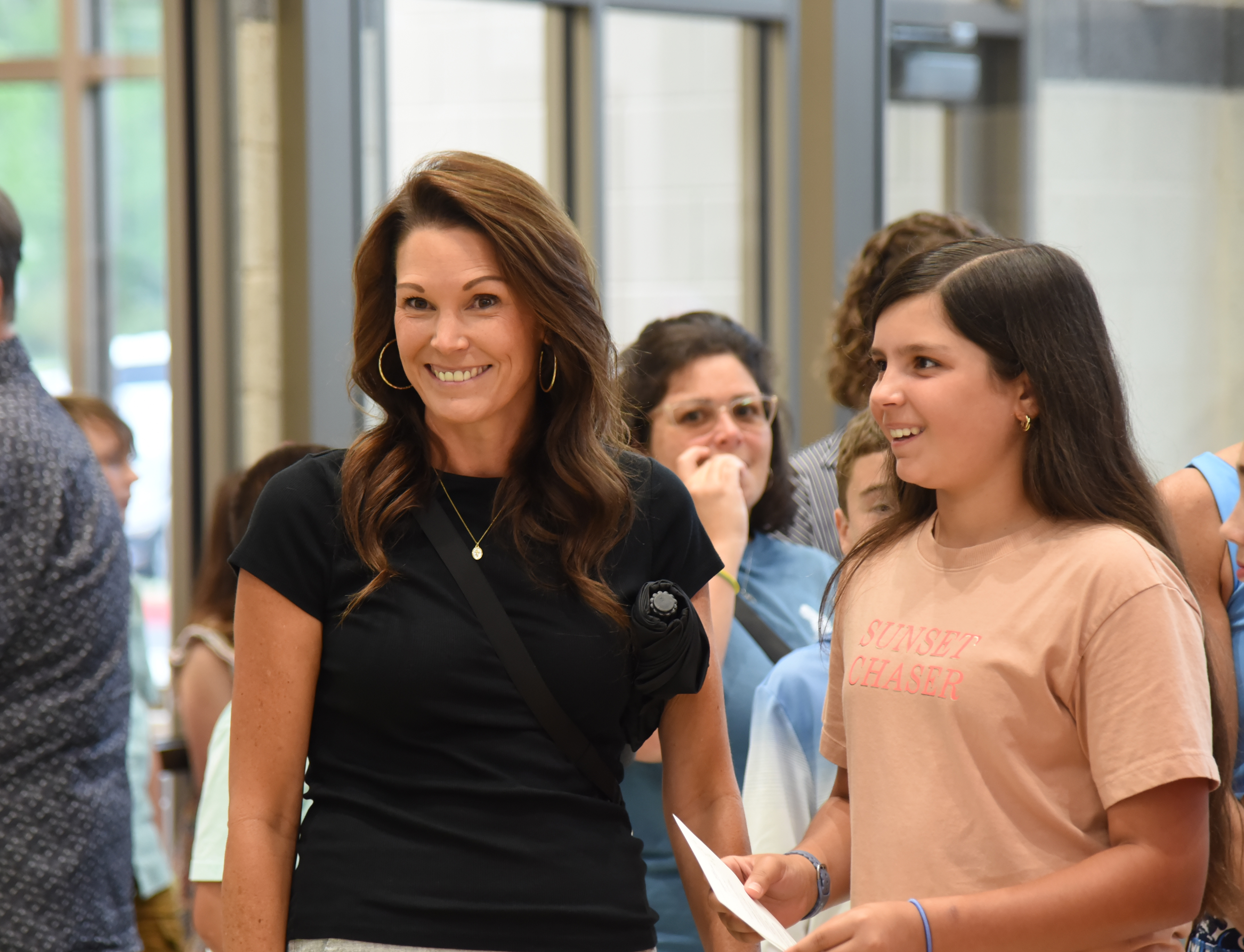 A smiling woman in a black top talks to a girl in a peach shirt in a busy hallway.