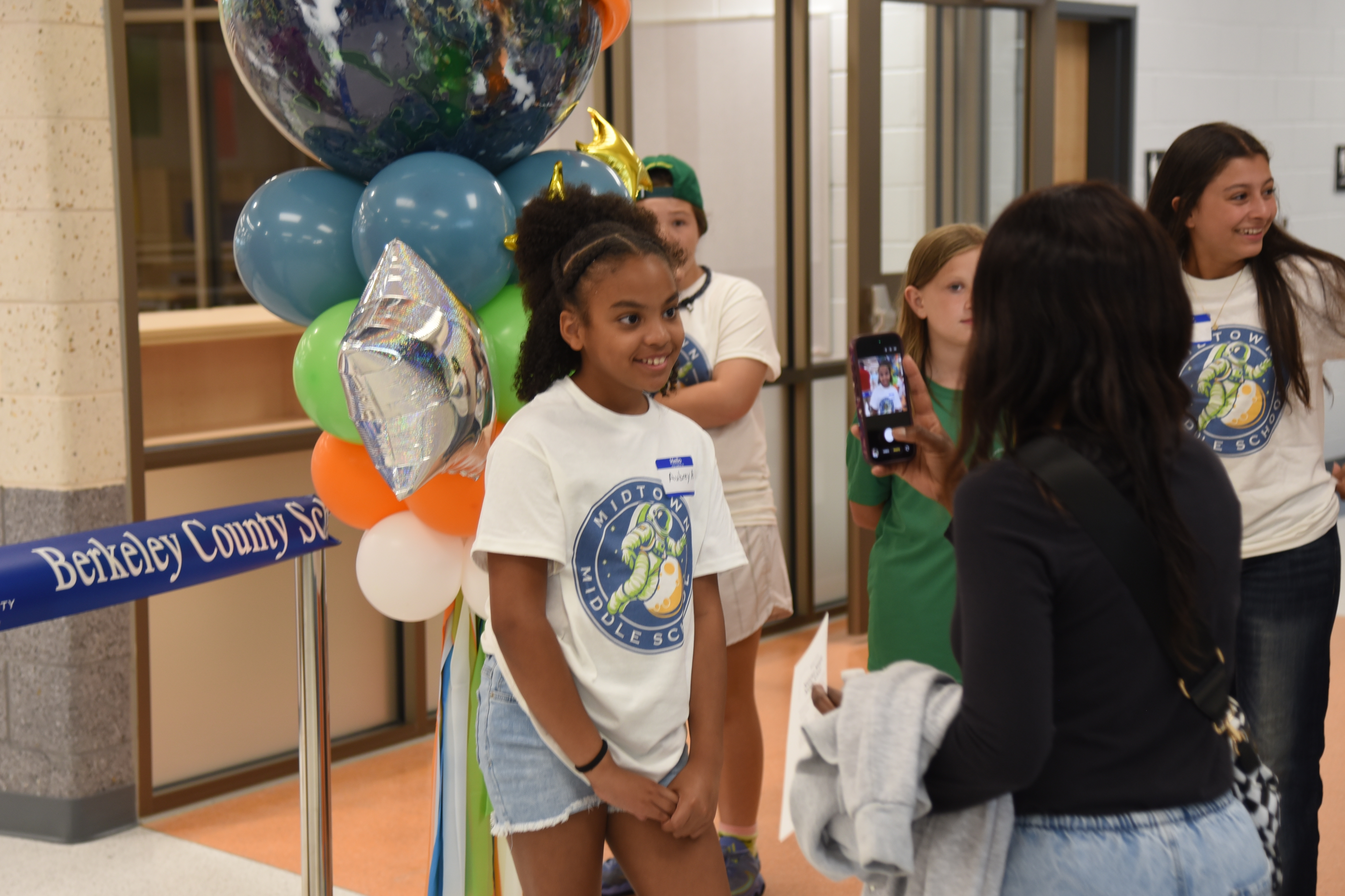Multiple people stand in a hallway. A young girl stands with balloons. A woman takes a photo.